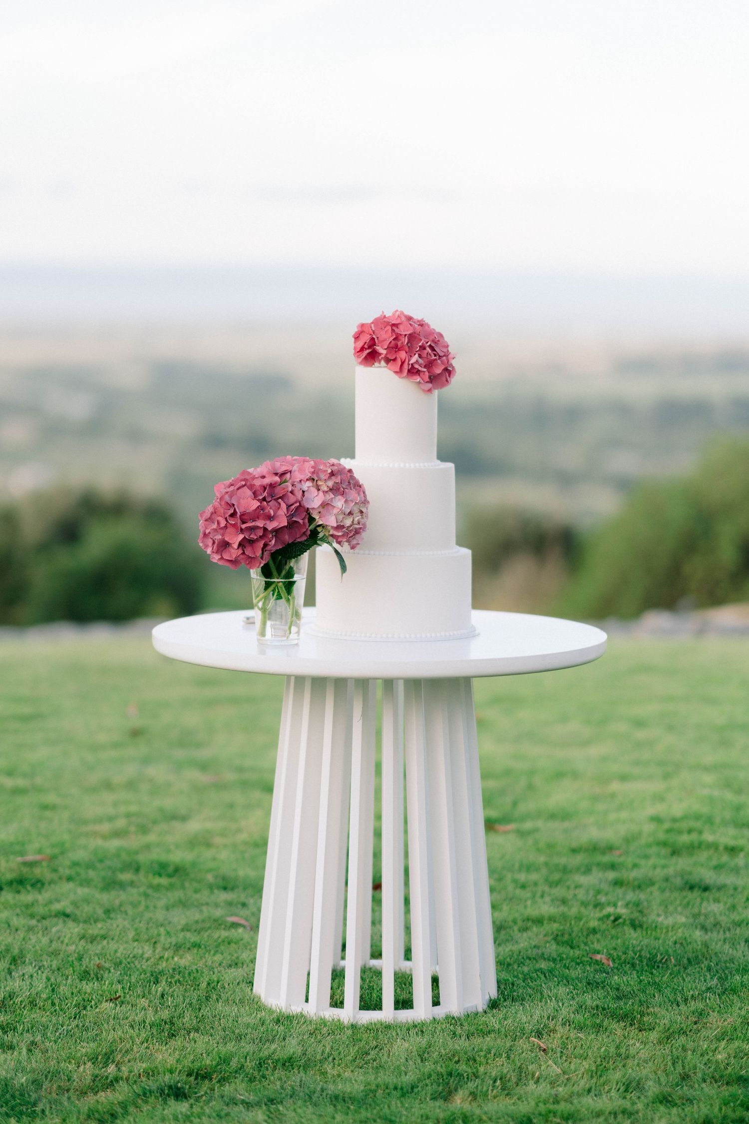 A modern white wedding cake decorated with bright pink flowers sits on a pedestal table outdoors.