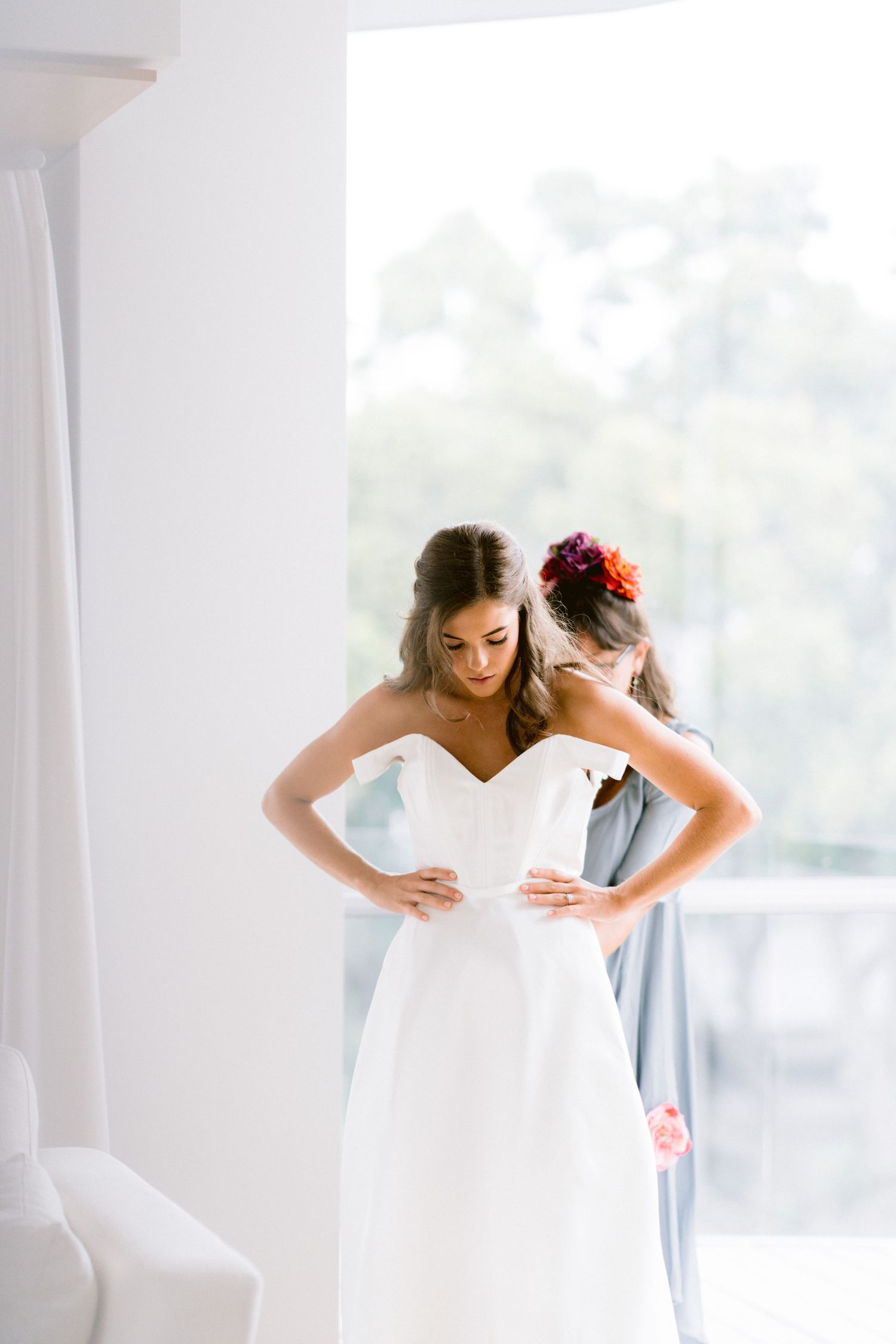 A bride gets dressed in a bright white wedding gown near sunlit windows.