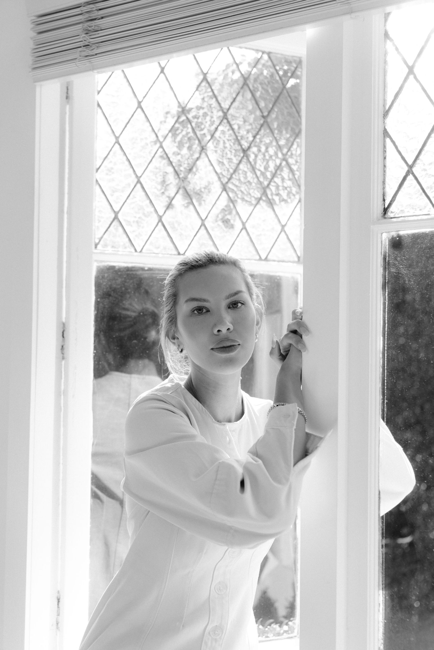 Black and white portrait of a woman in white clothing standing by a window with diamond pattern lattice.