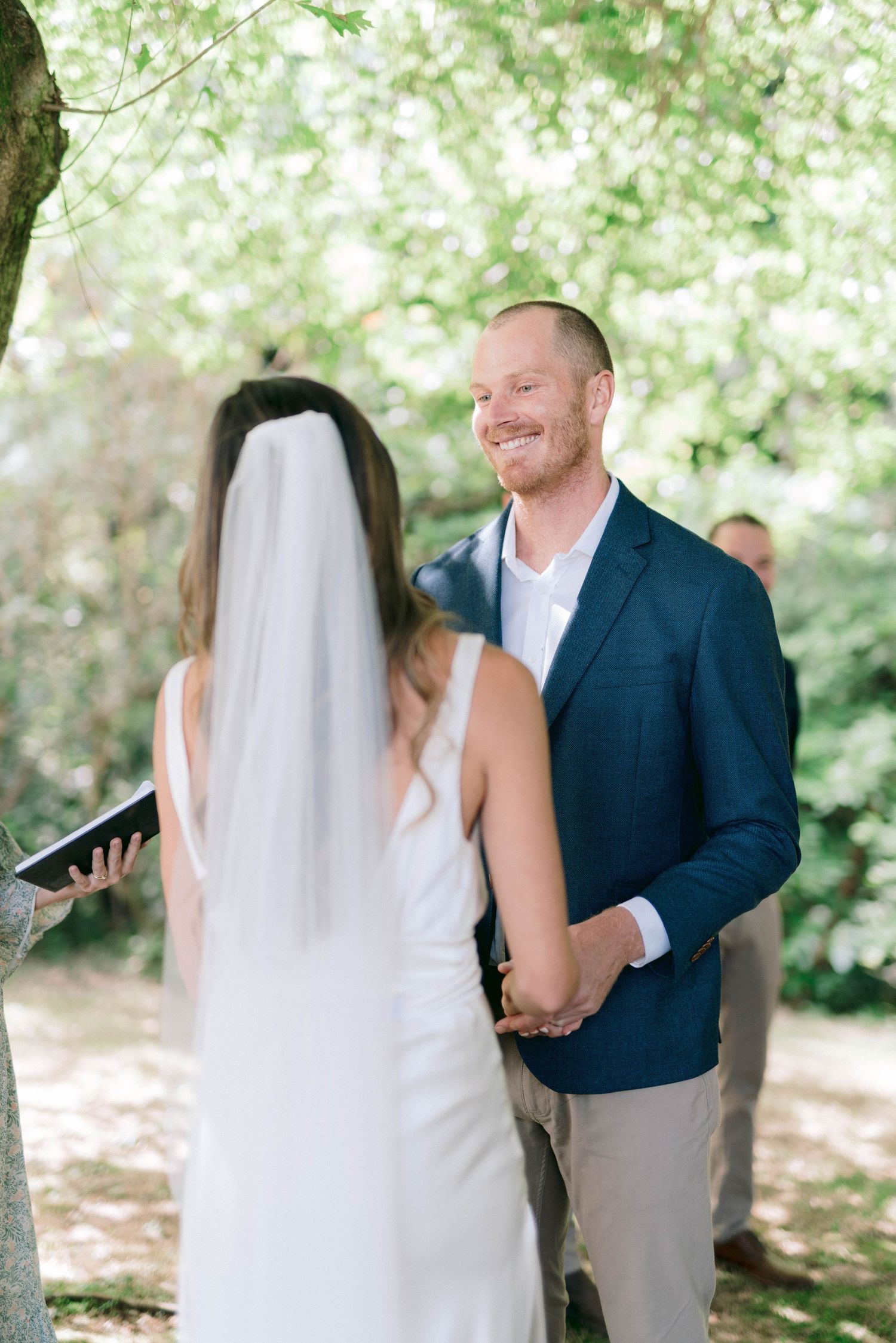 A wedding ceremony takes place outdoors under dappled sunlight with a couple exchanging vows.