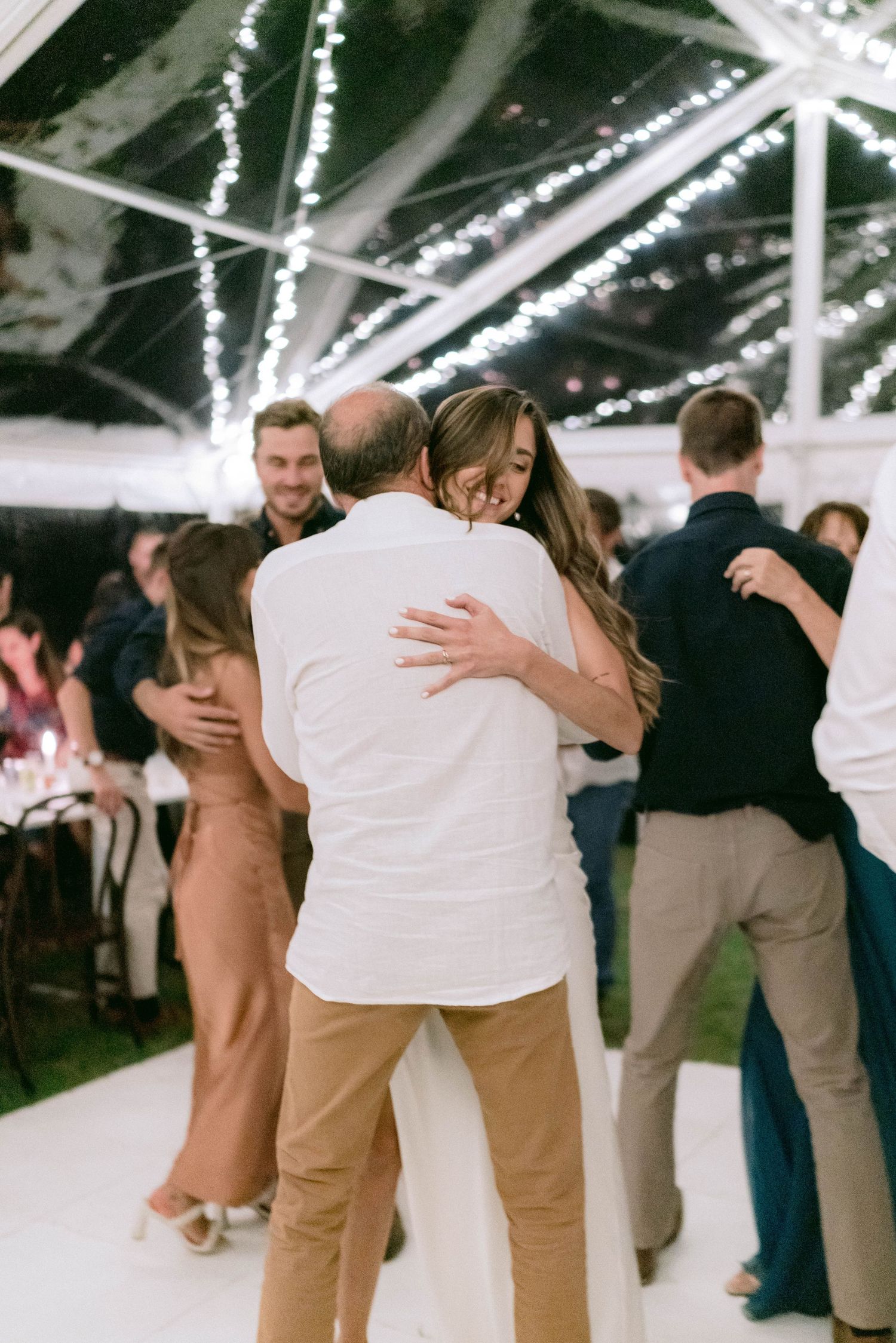Couples slow dance beneath a clear tent with twinkling lights at an evening wedding reception.