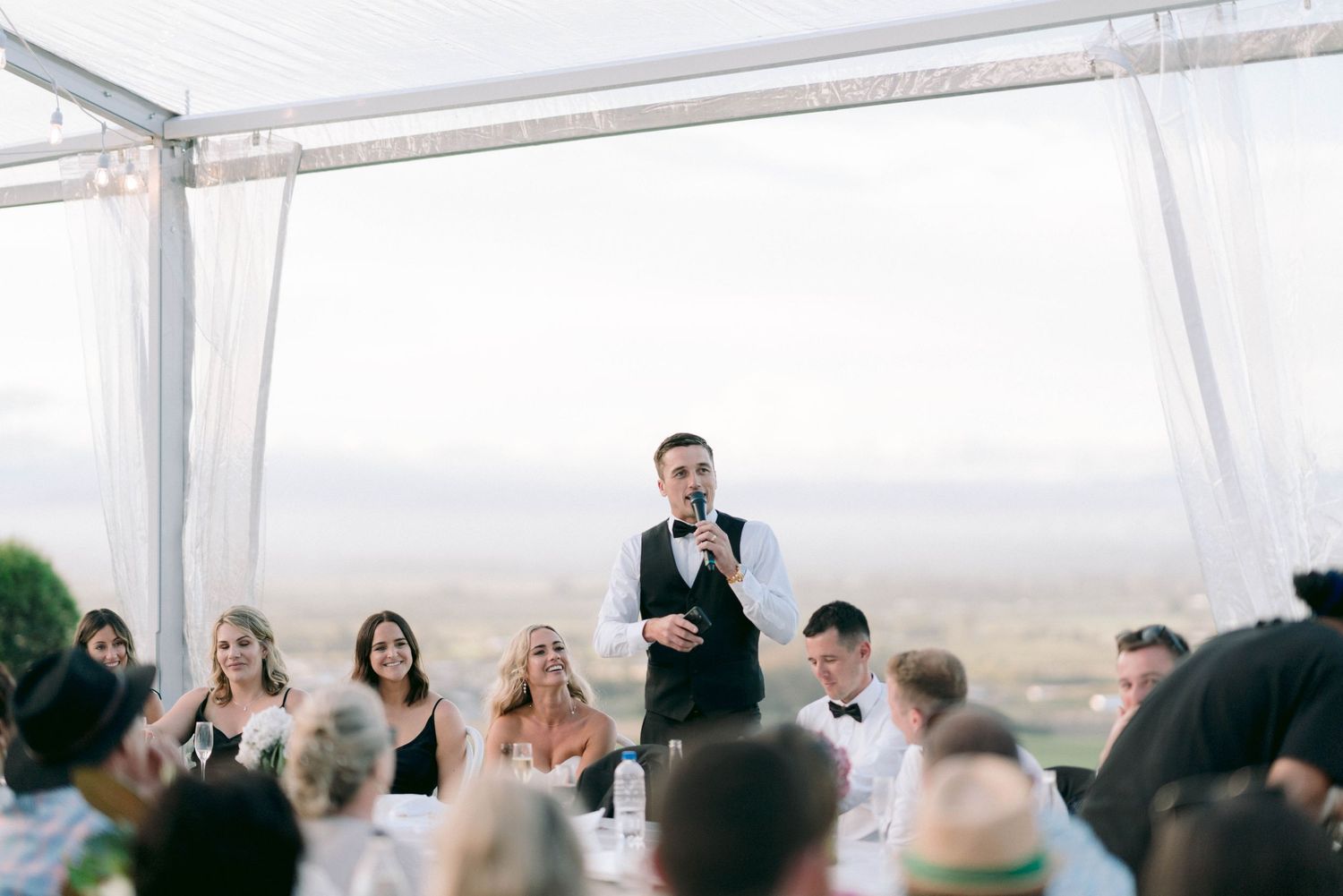 Wedding reception toast being given under a white tent with guests seated at tables.