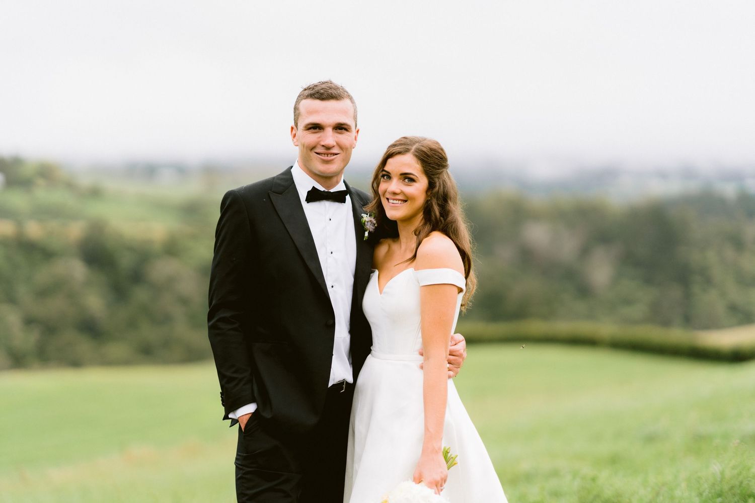 Couple poses for formal wedding portrait on a hilltop with scenic landscape view.