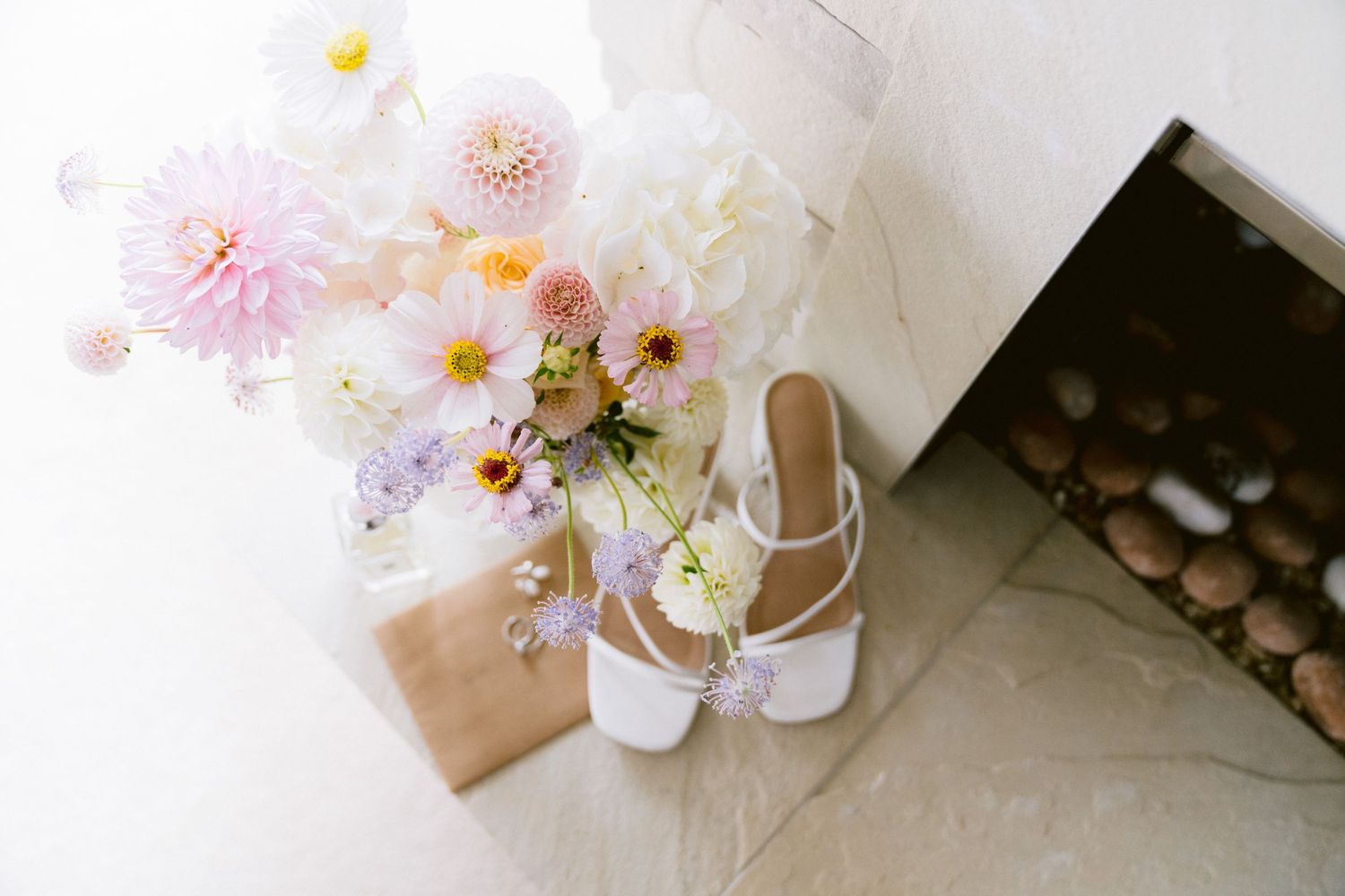 Two light pink dahlia flowers rest beside a white ceramic cup on a natural stone surface with scattered petals.