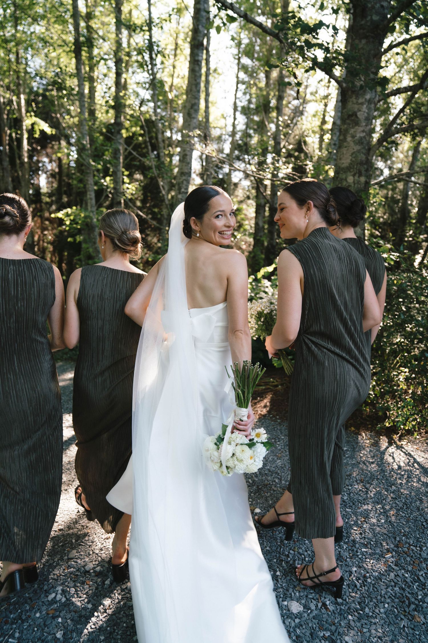 Bride in white gown looks back while walking with bridesmaids in sage green dresses through a sunlit forest path.