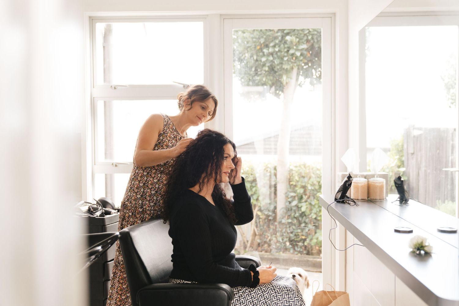 Two people in a bright sunlit room with white windows and kitchen counter space working together in a relaxed setting.