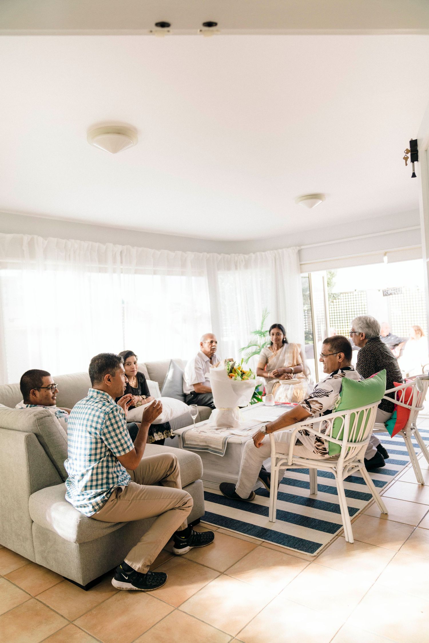 A group of friends gather in a bright sunlit living room for conversation and socializing around a coffee table.