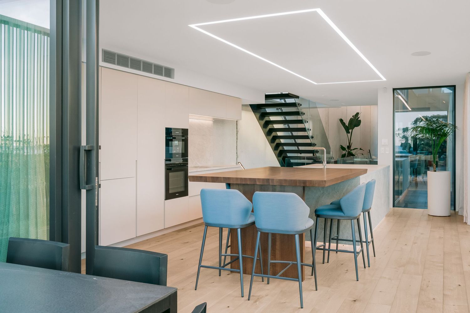 Modern dining area with blue bar stools and floating staircase under geometric ceiling light.