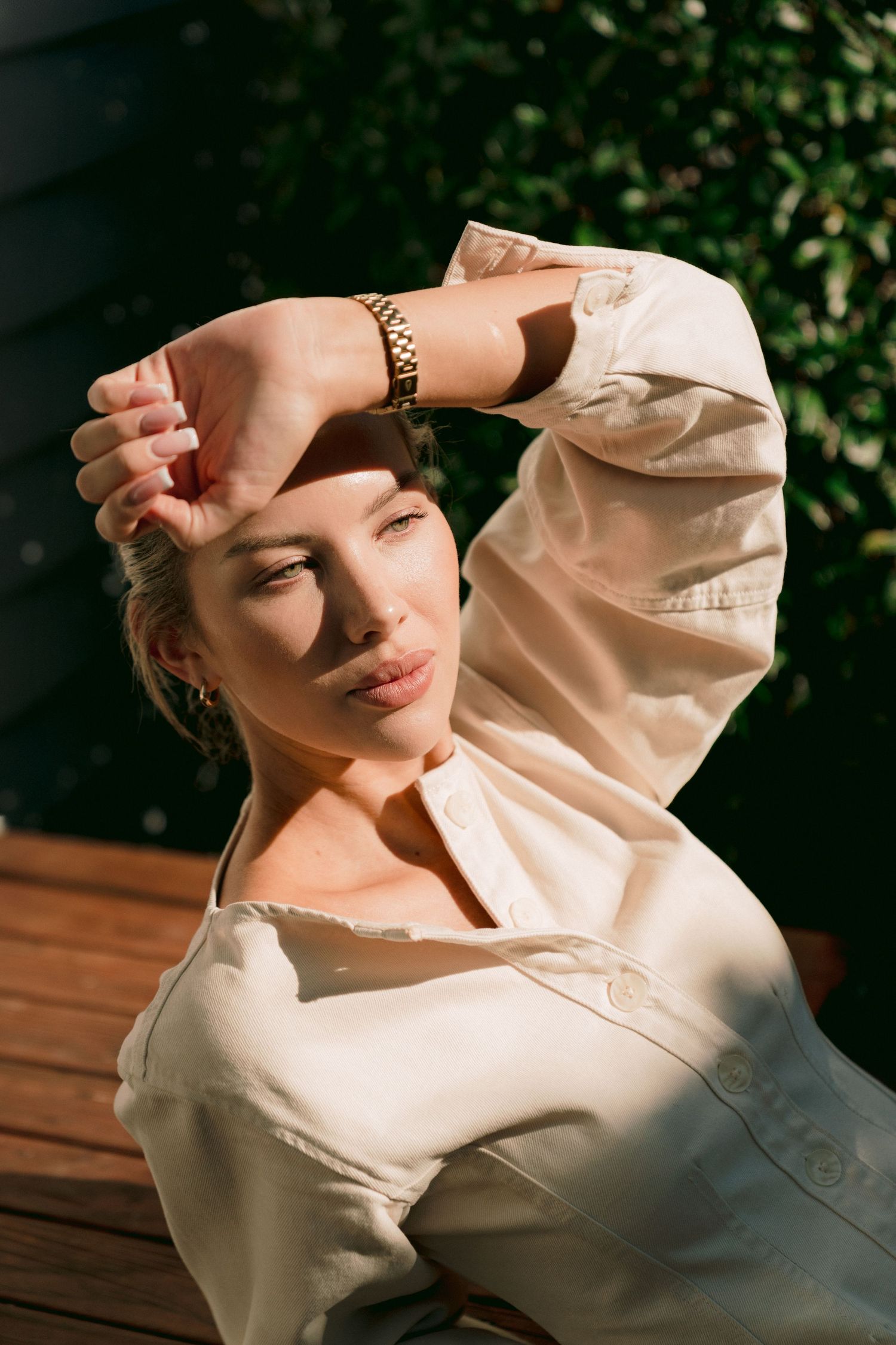 Person in cream colored blouse posing against greenery backdrop in warm natural sunlight.