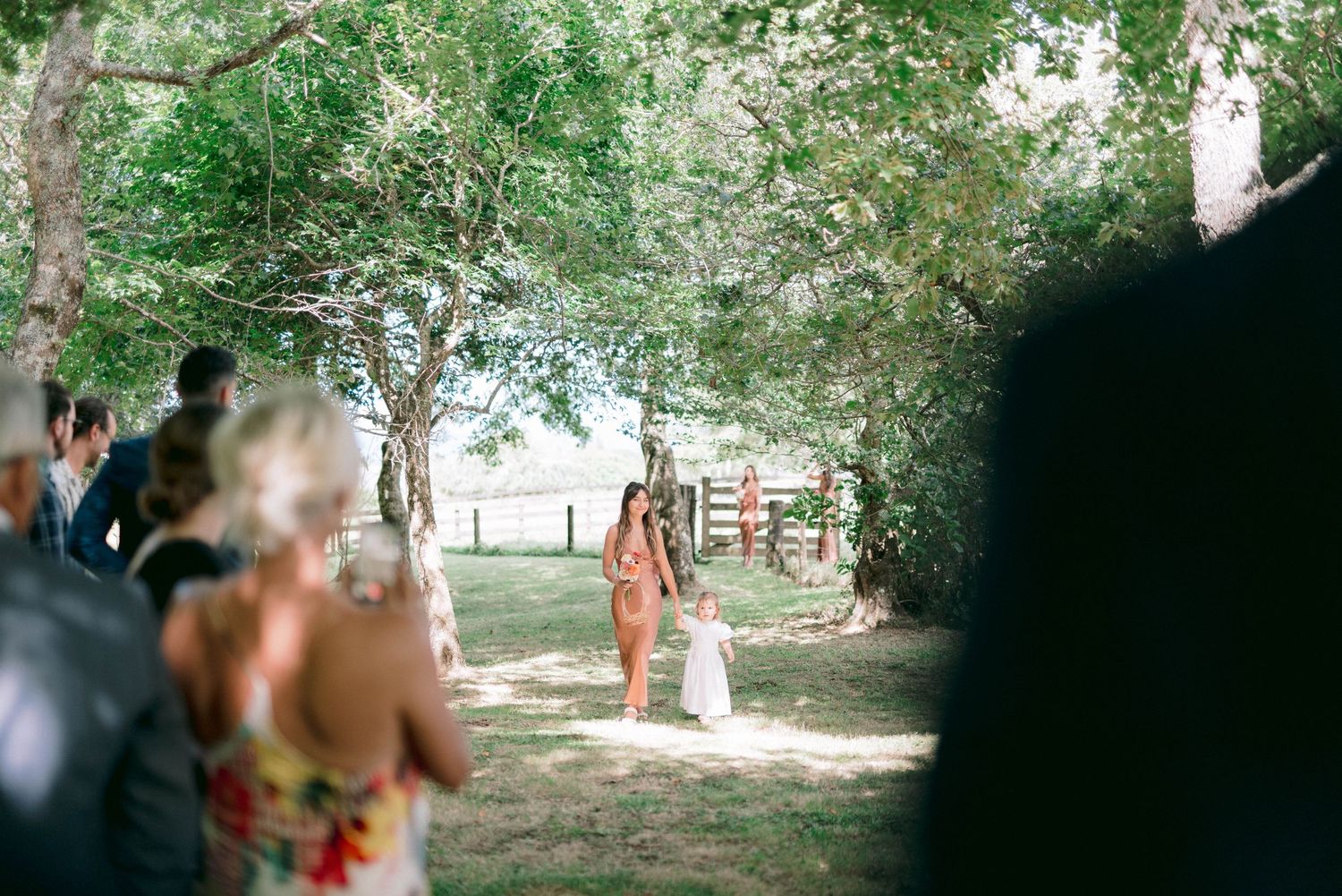 Wedding guests gather outdoors under trees at a rustic ceremony venue with white chairs set up on the lawn.