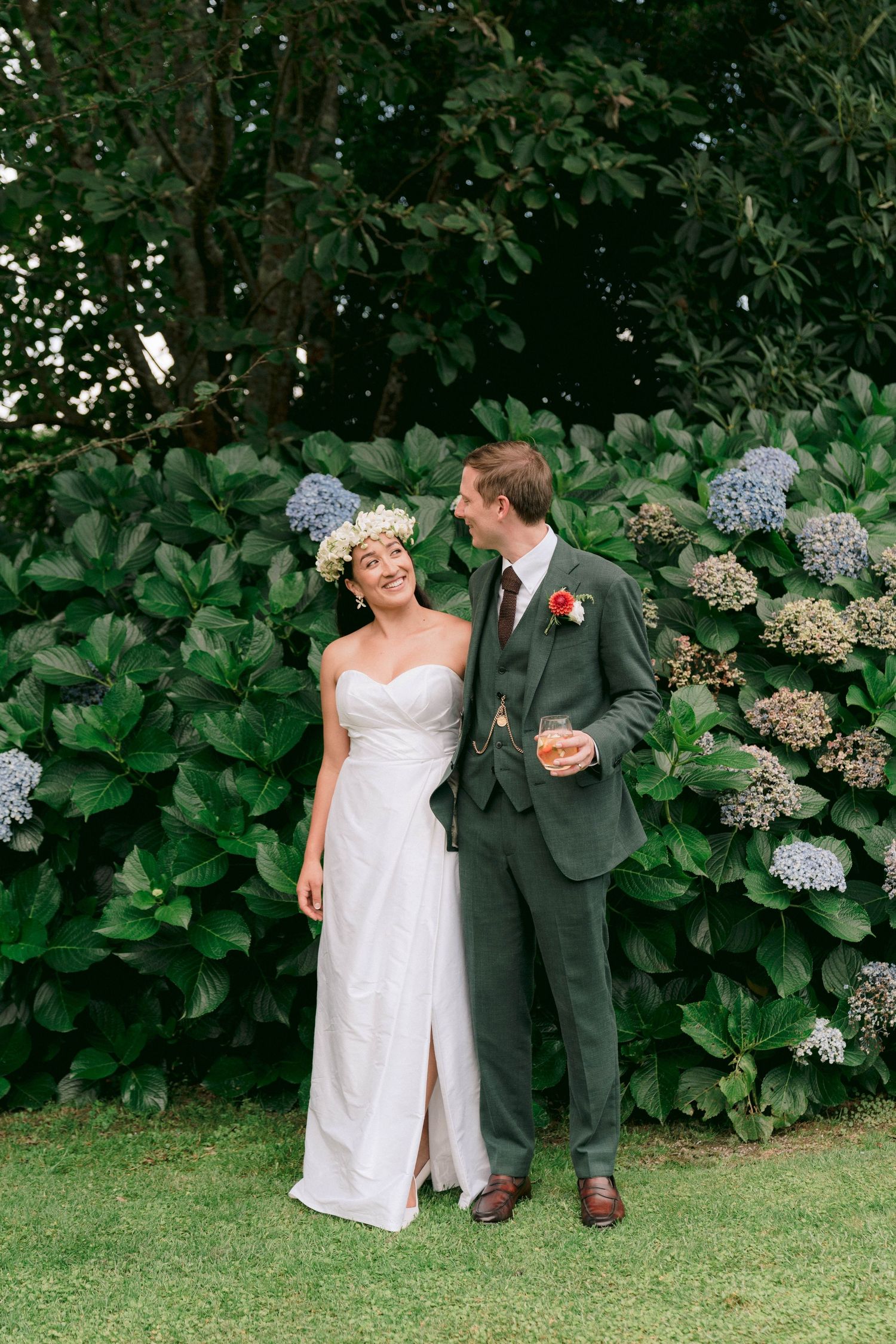 A romantic wedding couple poses in front of lush hydrangea bushes in a garden setting with beautiful greenery.