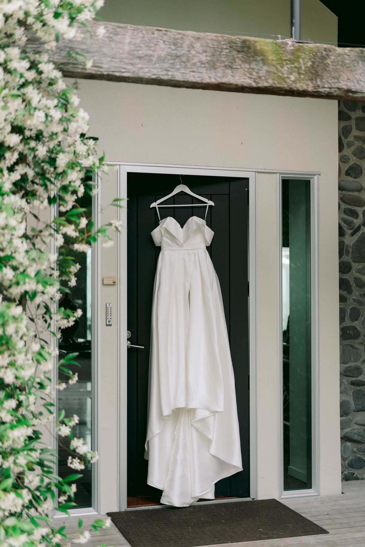 Elegant white wedding dress hanging on display against a dark door with blooming flowers nearby.