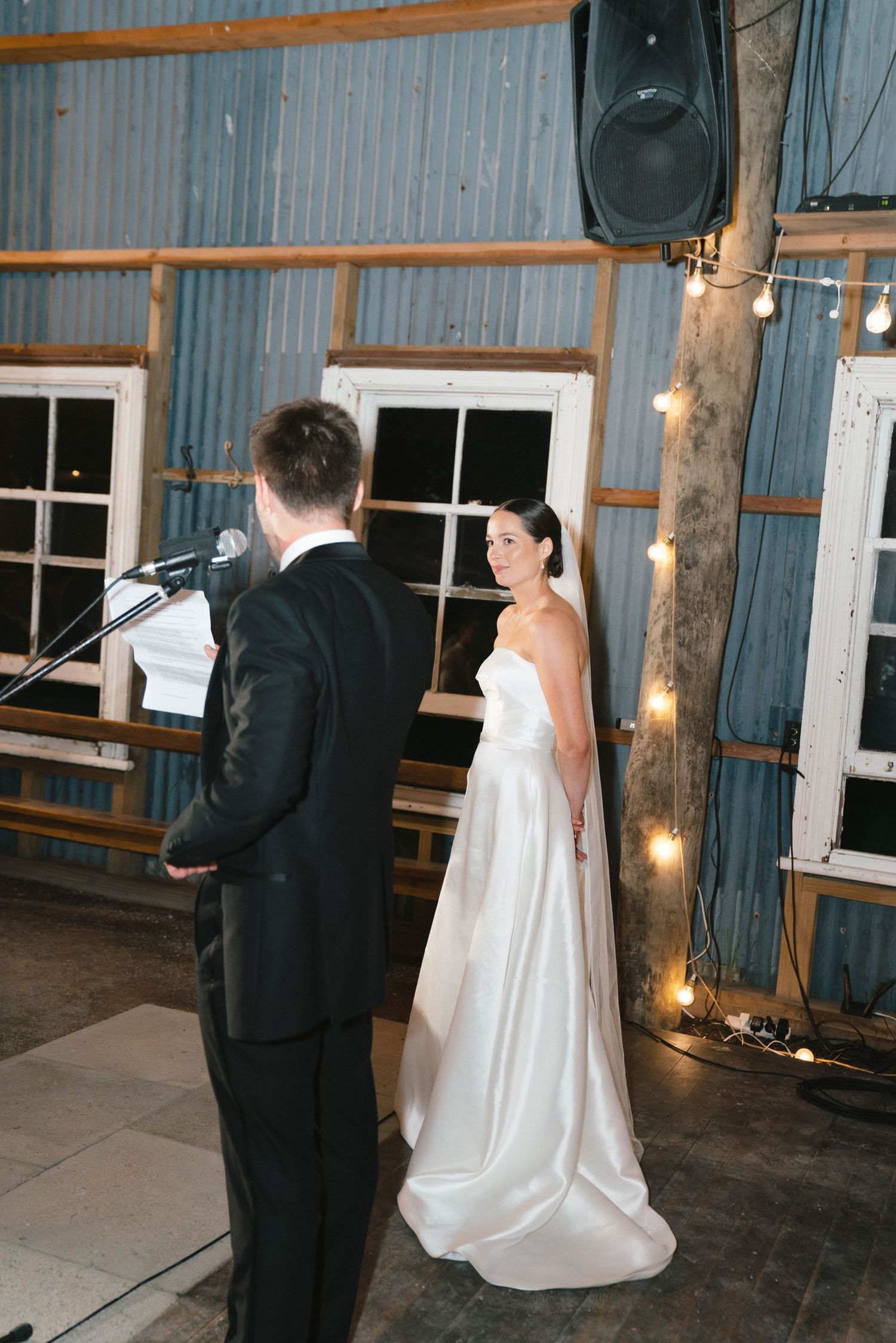 Bride in elegant white dress standing beside groom during wedding ceremony in rustic barn venue.