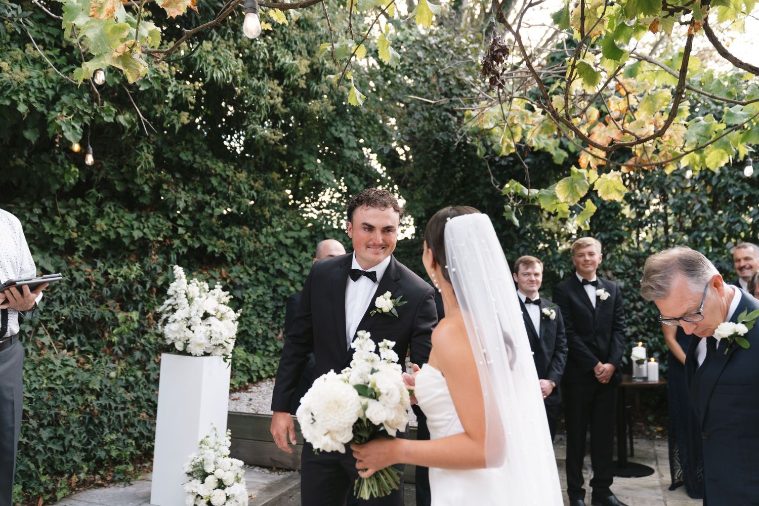 Newlyweds stand at outdoor wedding ceremony with white floral decor and greenery in garden setting.
