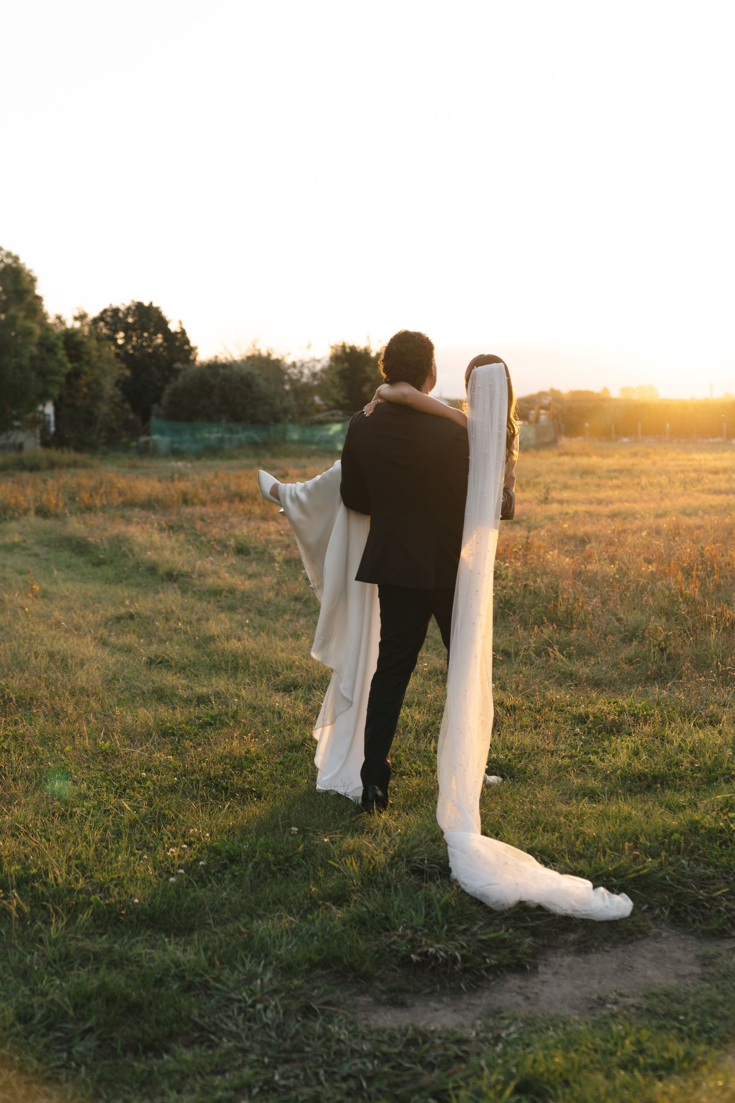 Wedding dress and black suit photographed against a golden sunset in a grassy field.