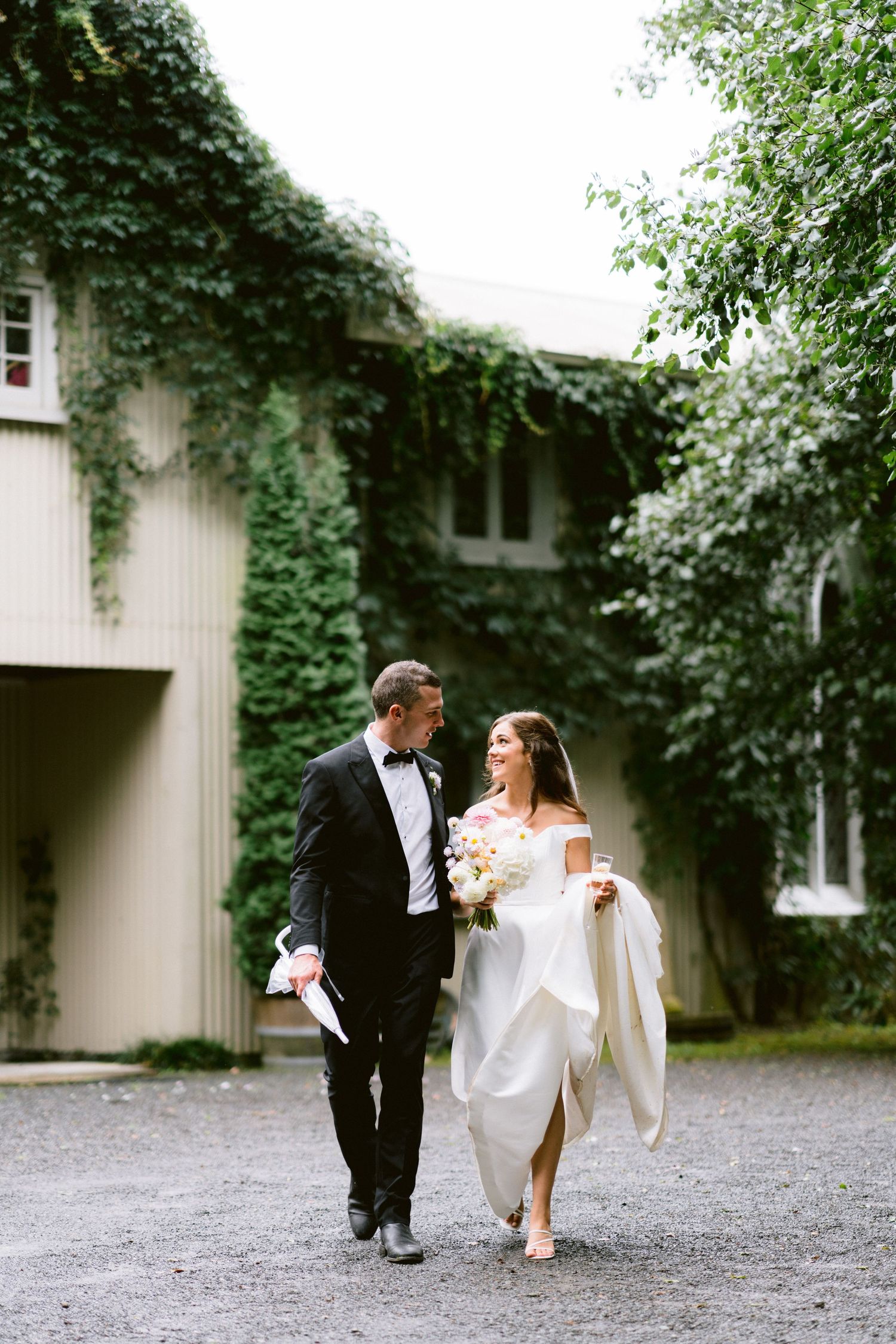 Newlyweds walk together on a gravel path beside an ivy-covered building.