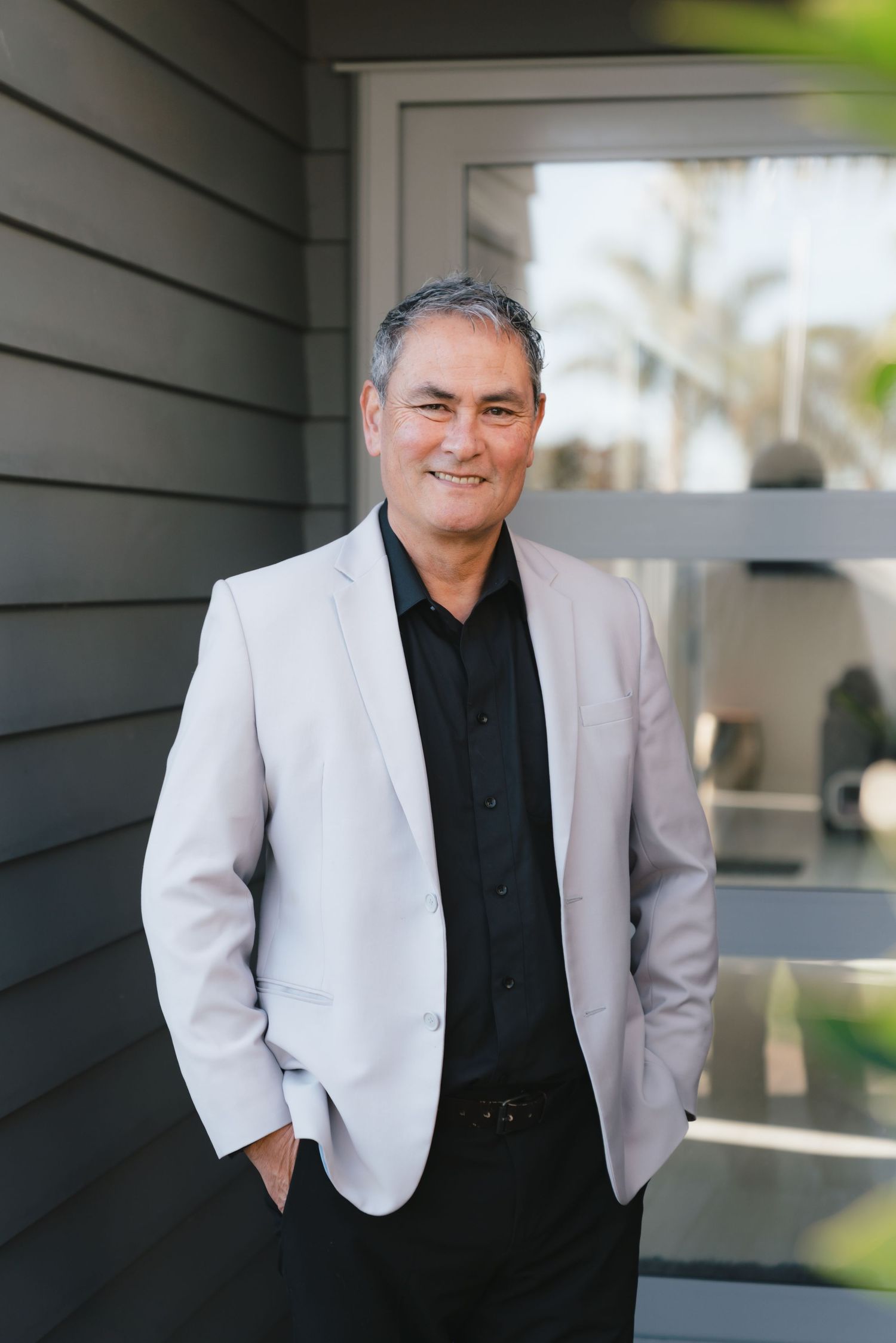 A person in a white blazer and black shirt poses against a gray exterior wall during a photoshoot.