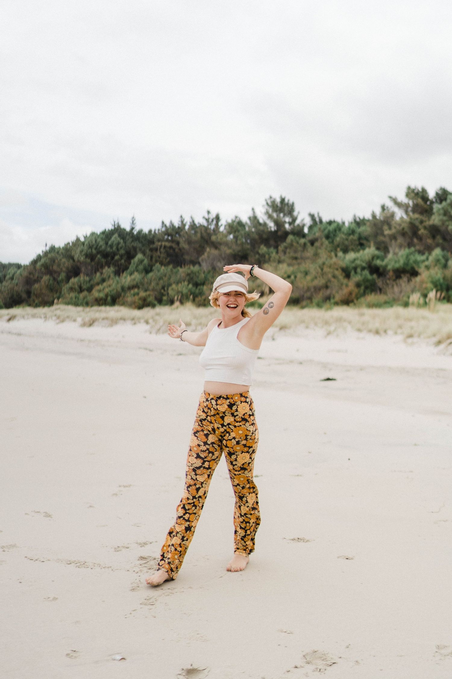 Person dancing joyfully on beach wearing floral pants and white tank top.