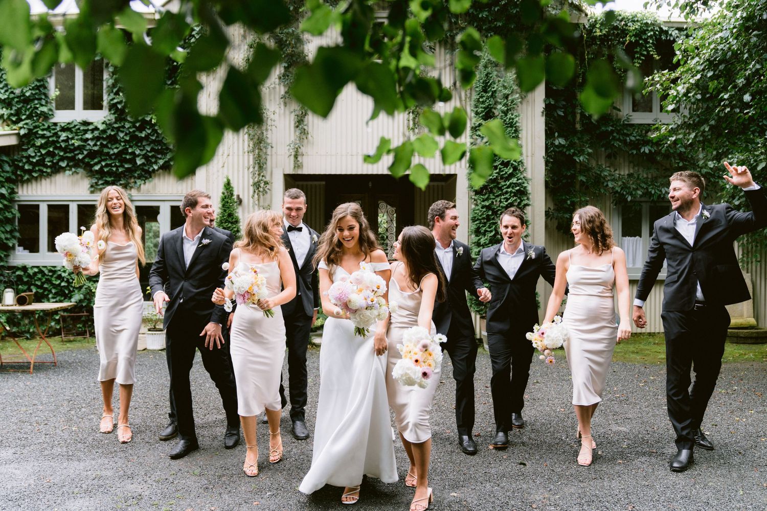 Wedding party walking together on a tree-lined path outside a stately building.