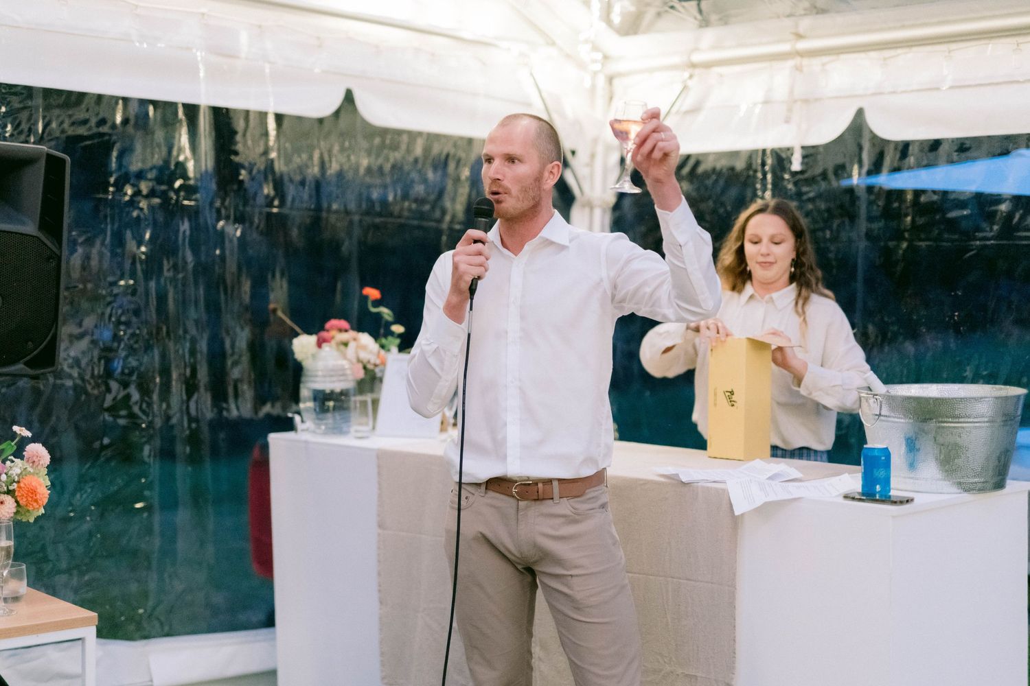 People making a speech at an outdoor event under a tent with white decor and tablescapes.