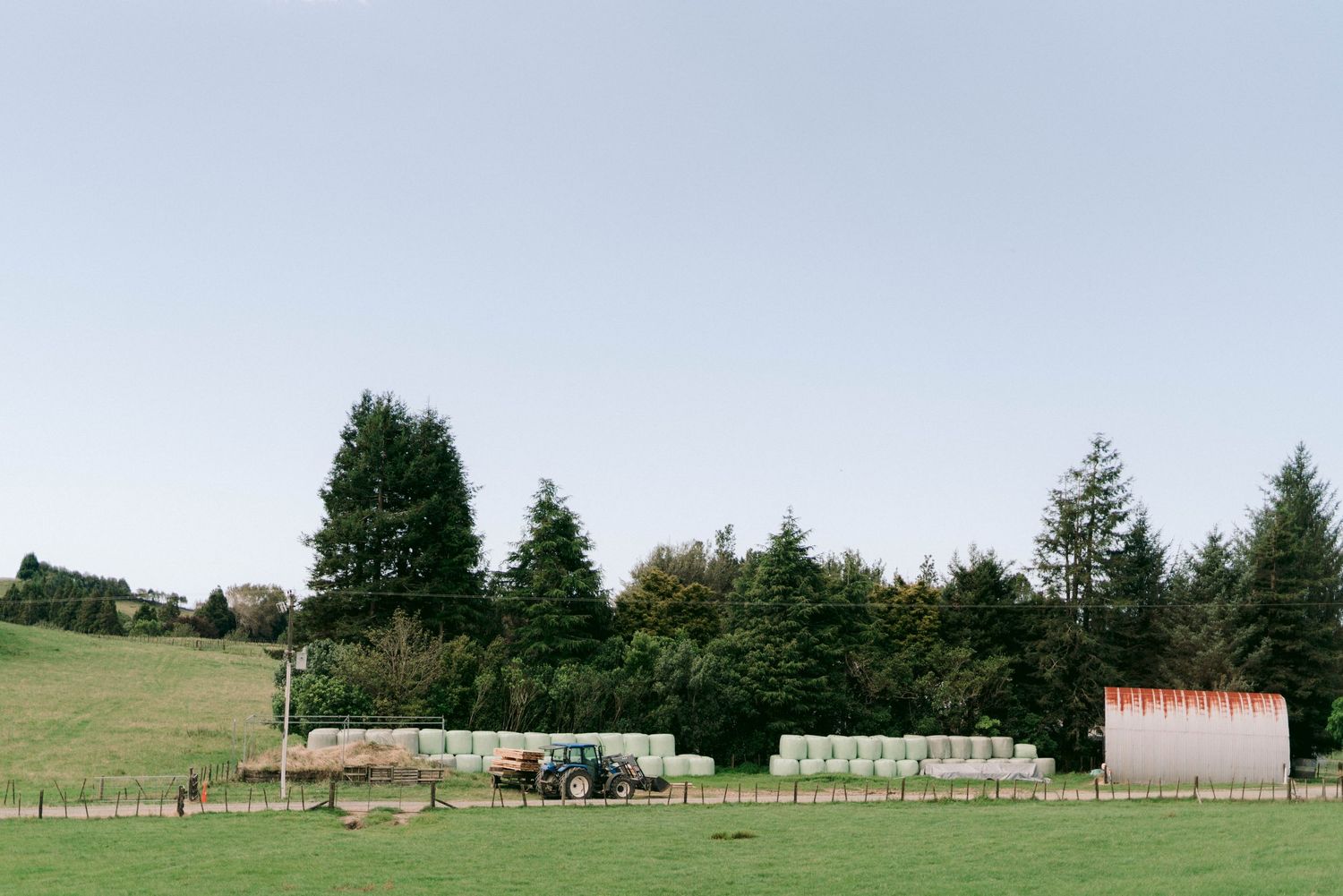 Rural farm setting with tractors and storage buildings against a backdrop of evergreen trees.
