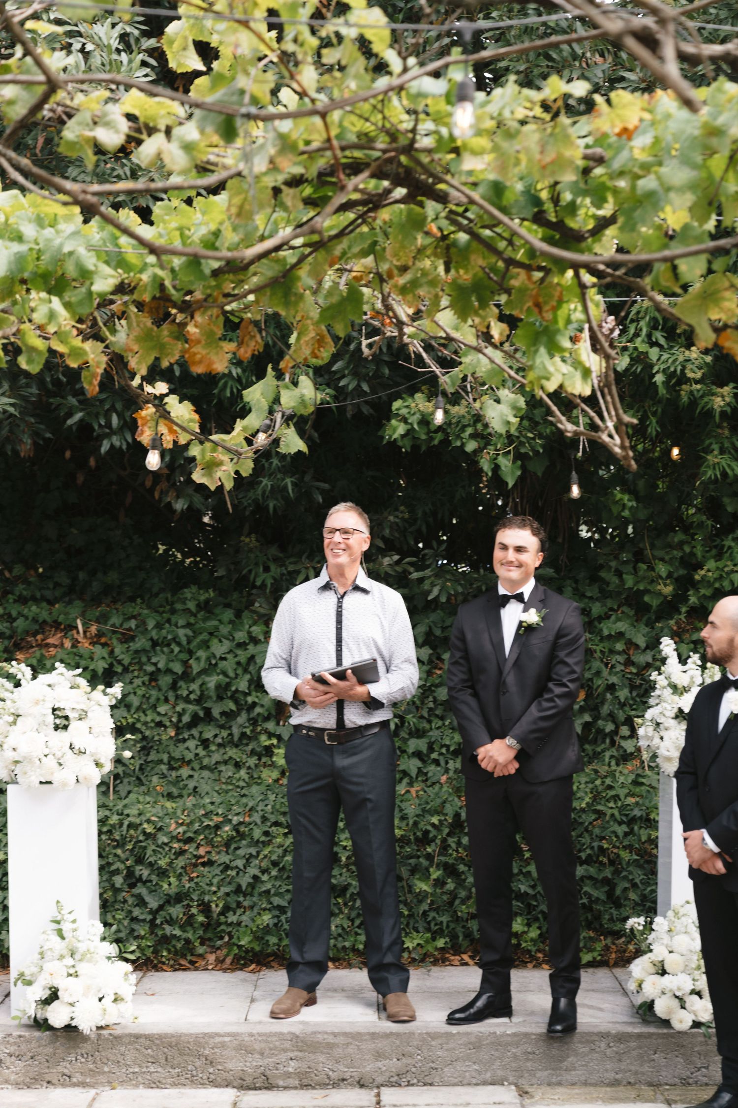 Wedding party members in formal attire stand along a garden wall with white floral decorations.
