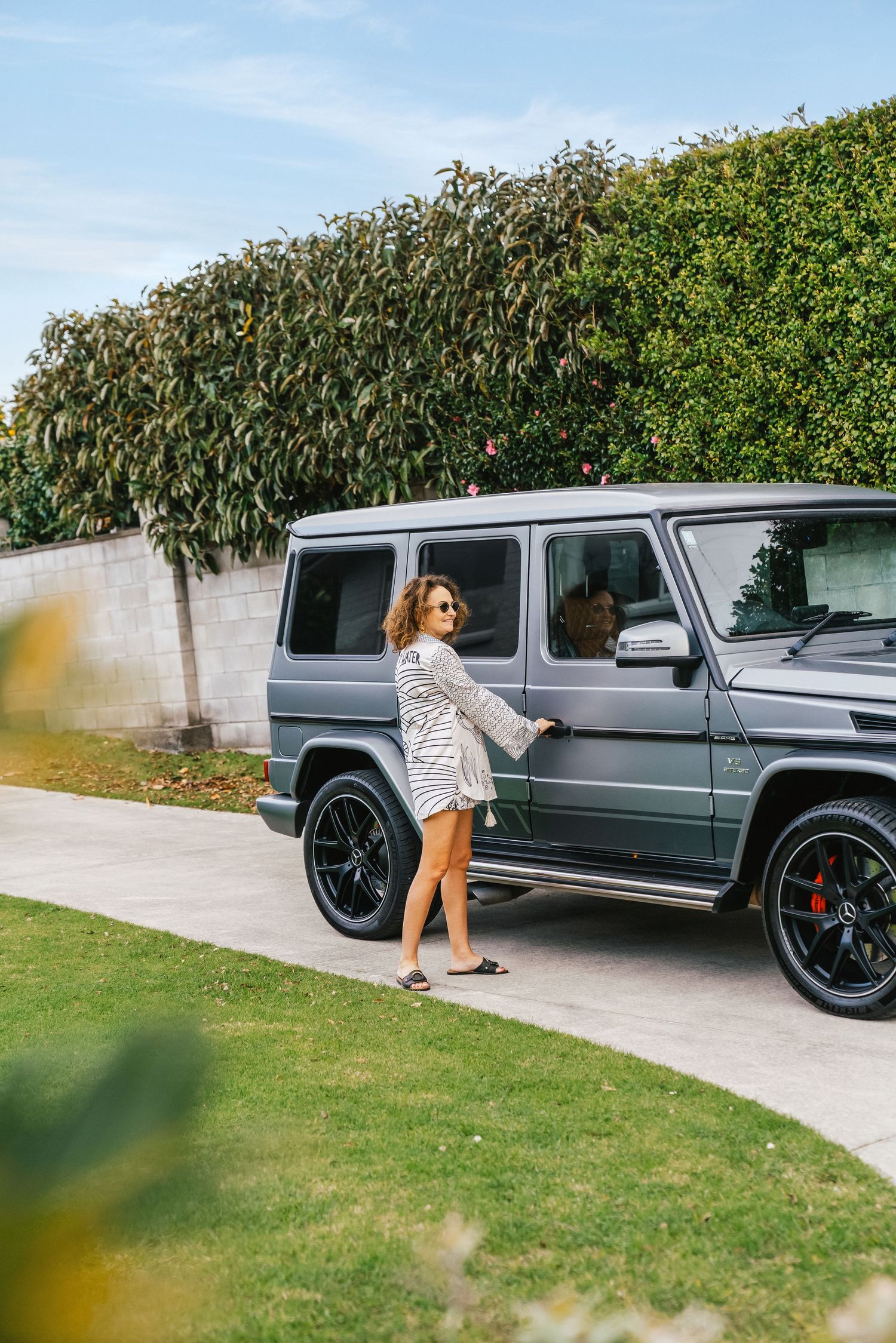 Person in striped dress stepping into a silver Mercedes G-Class SUV parked on a driveway next to lush greenery.