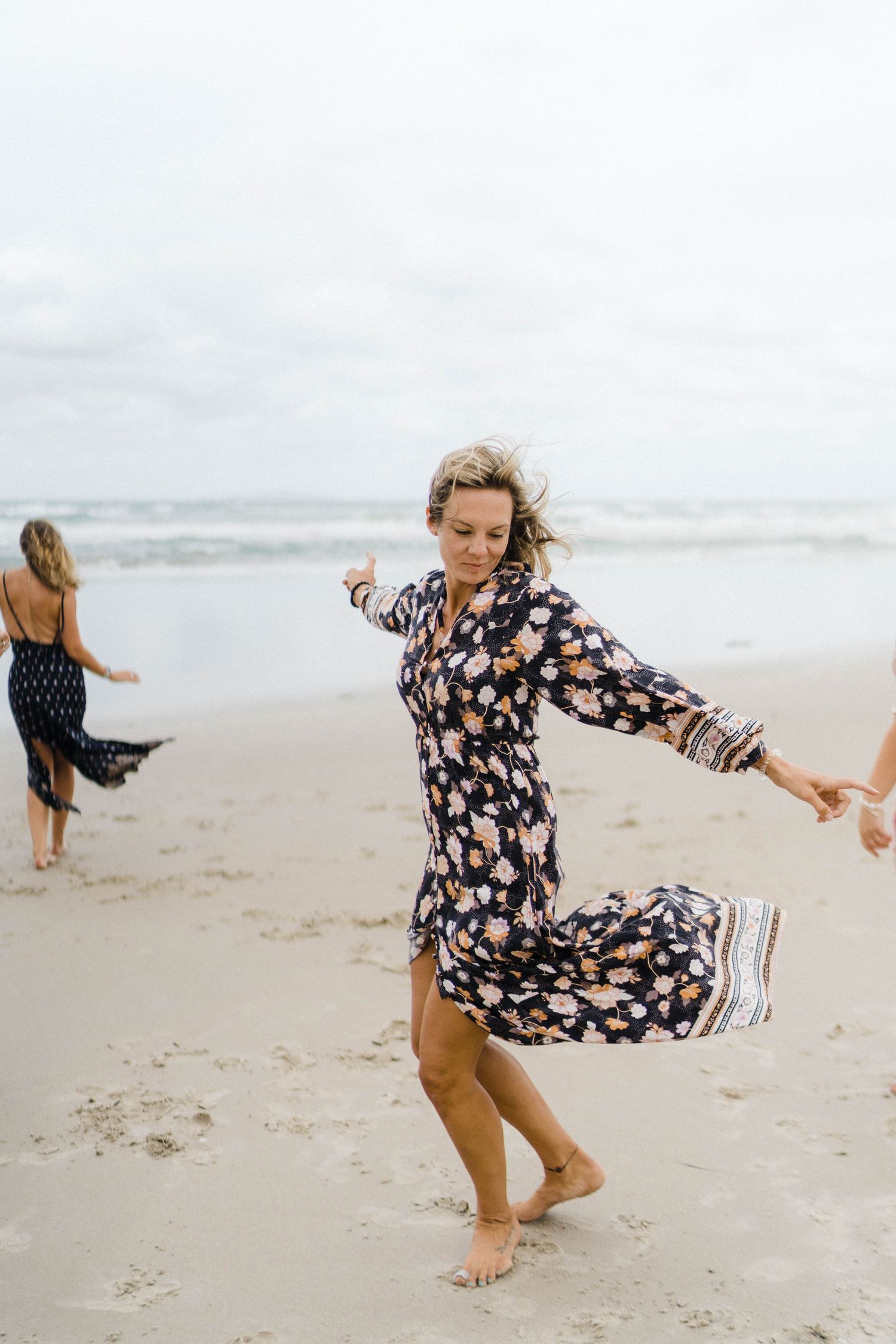Someone in a floral print dress twirls playfully on a sandy beach with waves in the background.
