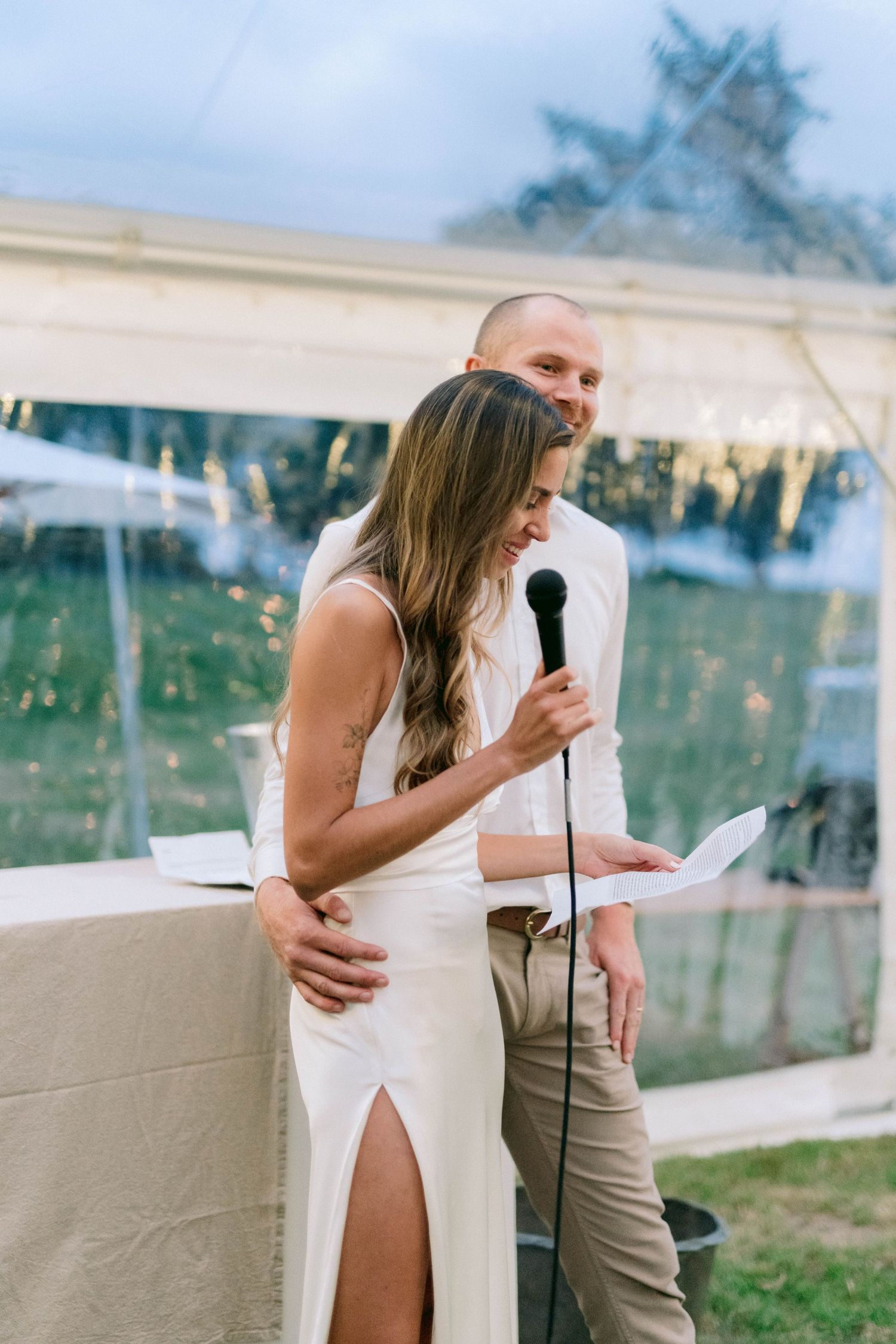 Couple sharing an intimate moment during wedding speeches under an outdoor tent.