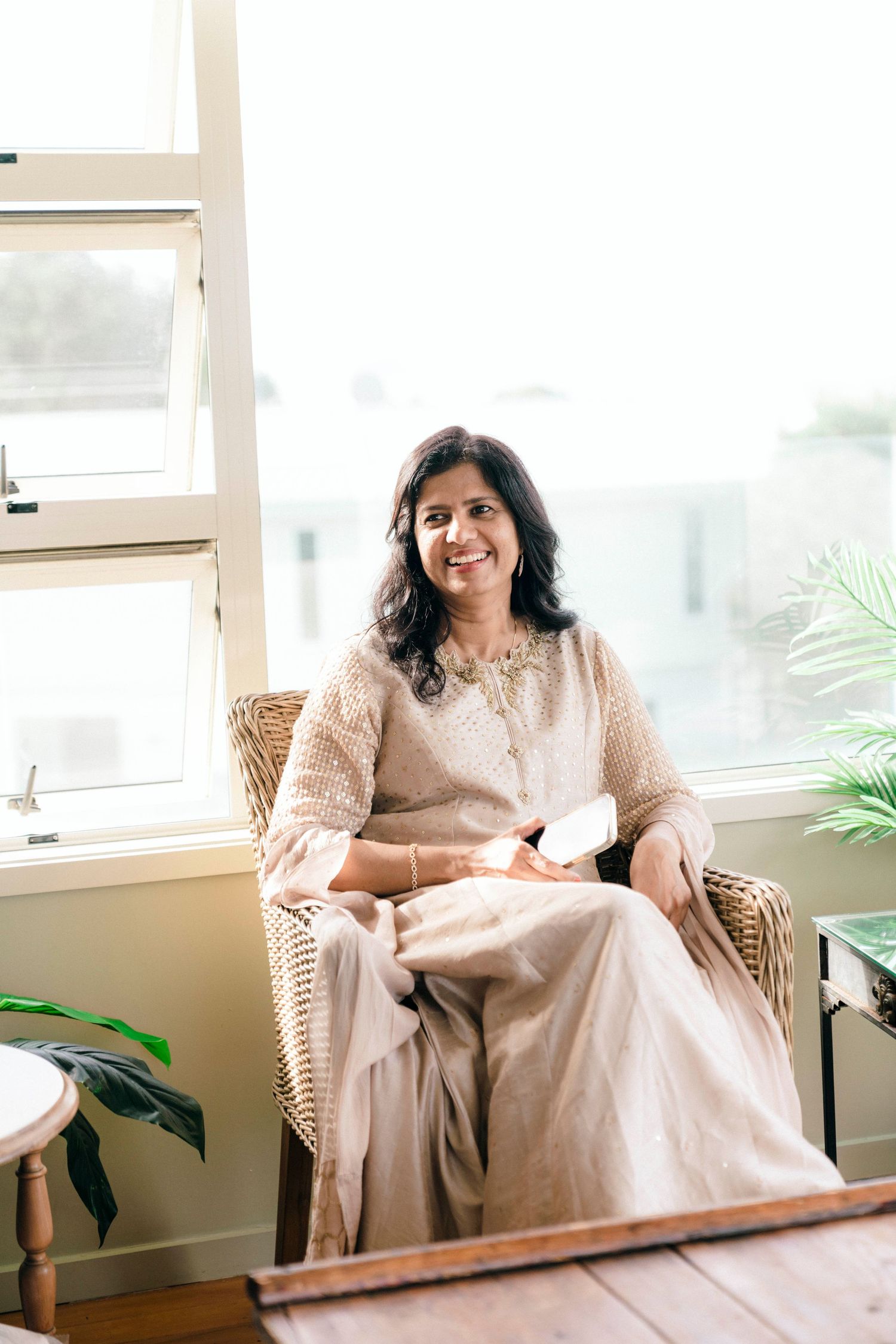 A person in a beige outfit sits relaxed in a wicker chair by a bright window with indoor plants nearby.
