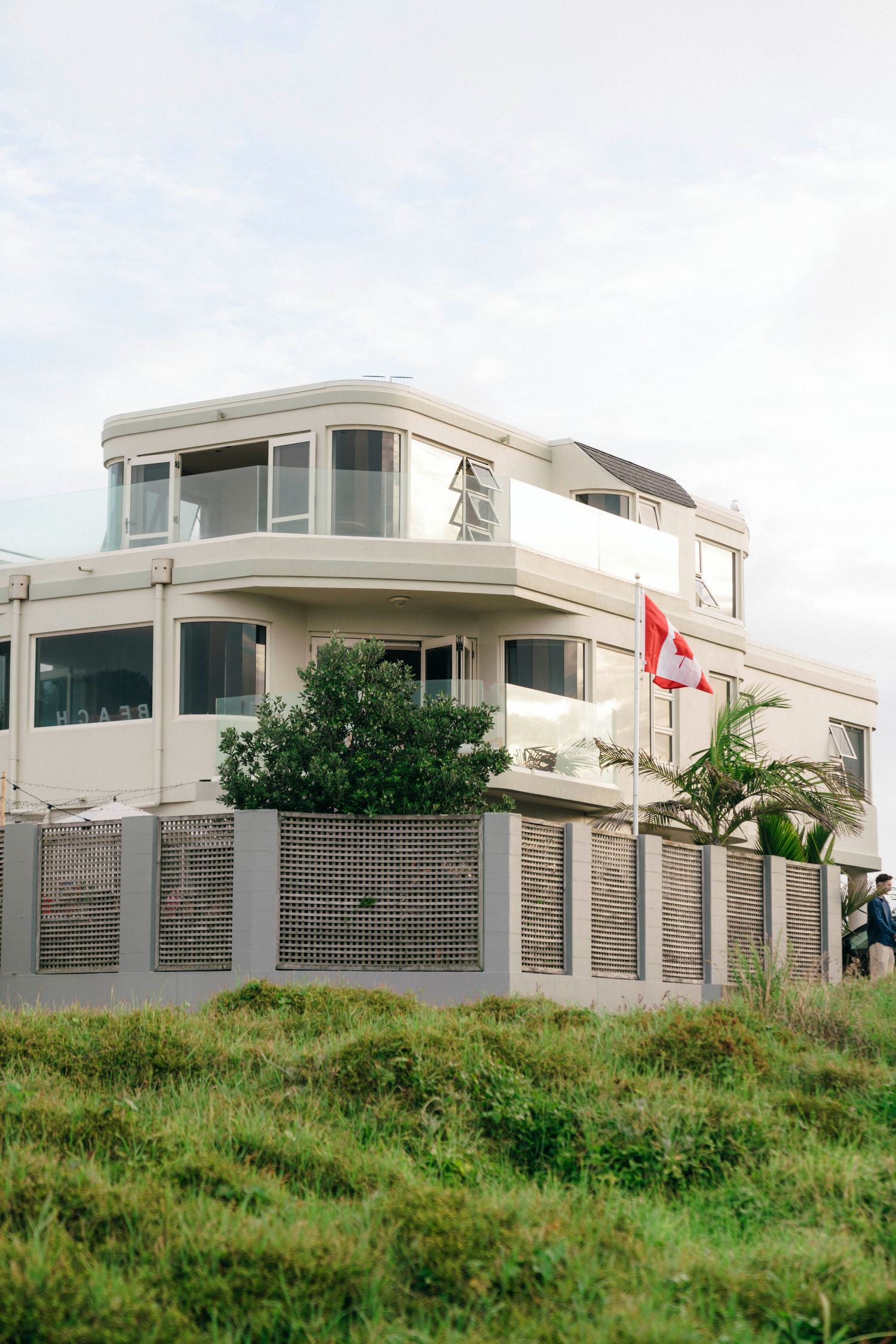 Modern white coastal house with glass windows and a red flag features metal fencing and lush greenery on a grassy slope.