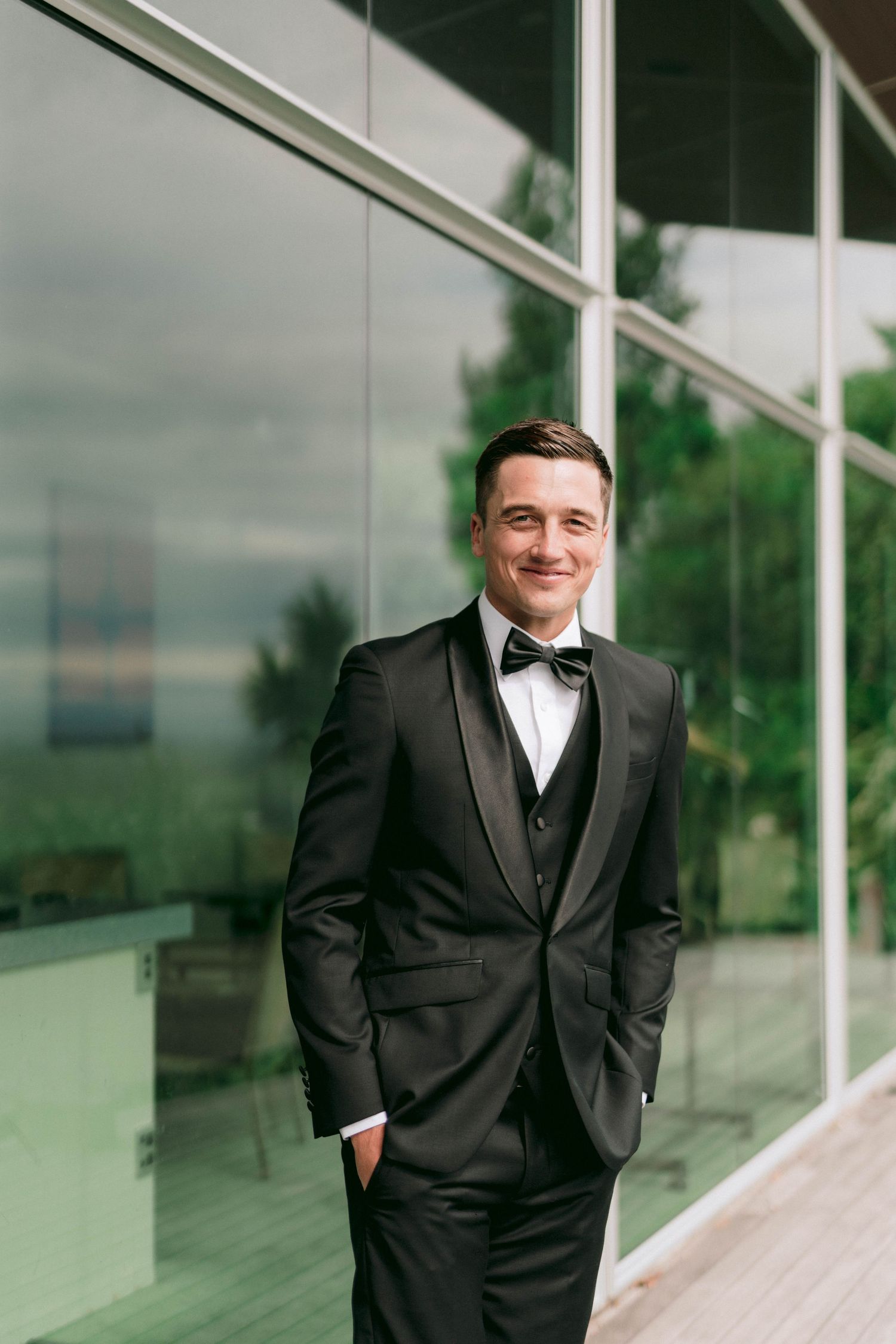 Formal portrait of person in black tuxedo standing against modern glass building.