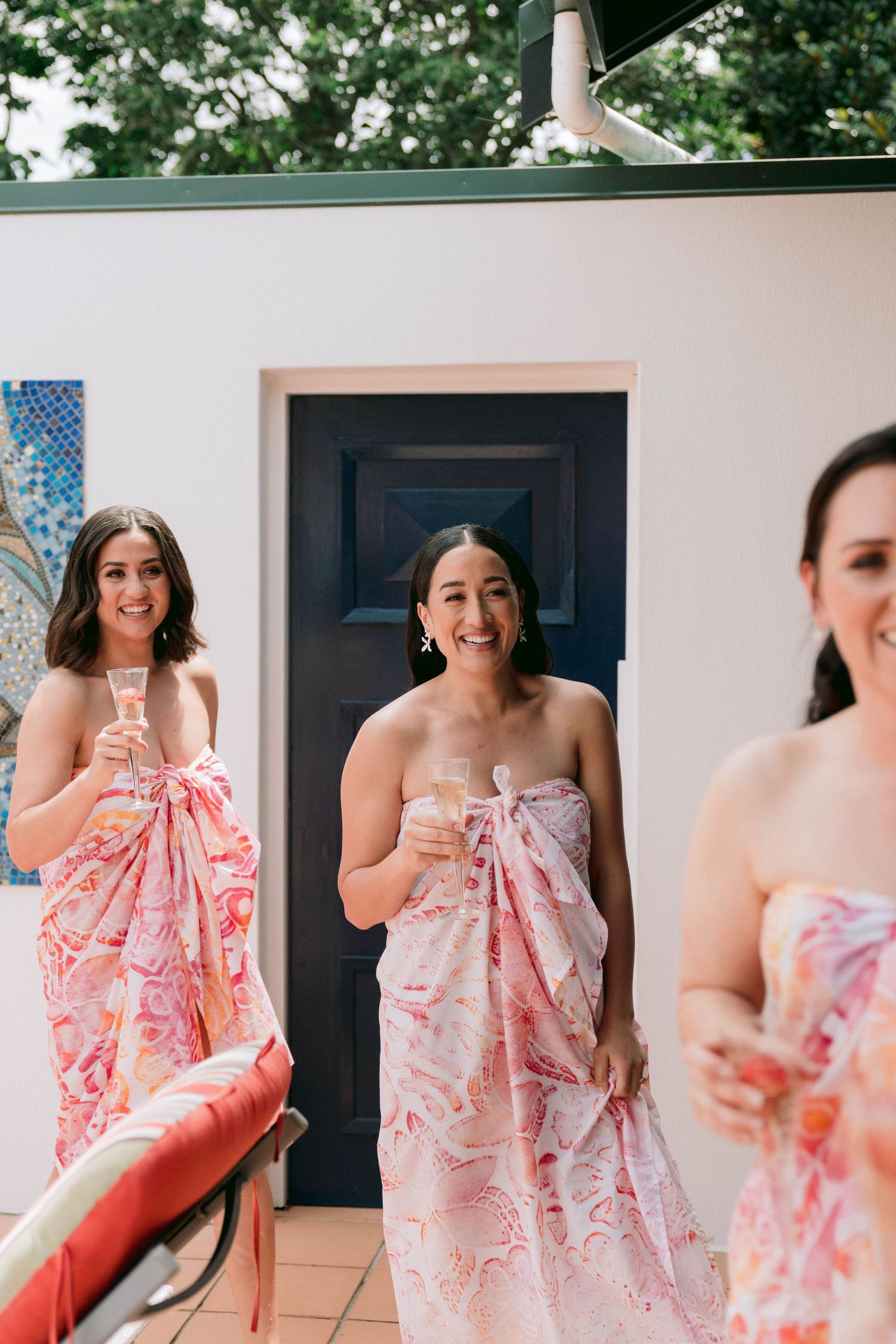 Wedding party members wearing pink floral print wraps stand together on a patio with champagne glasses.