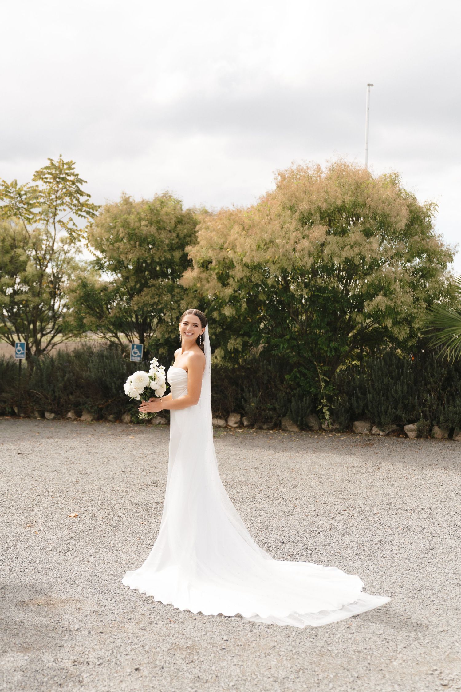 Bride in elegant white wedding dress with long train poses outdoors holding a white floral bouquet.