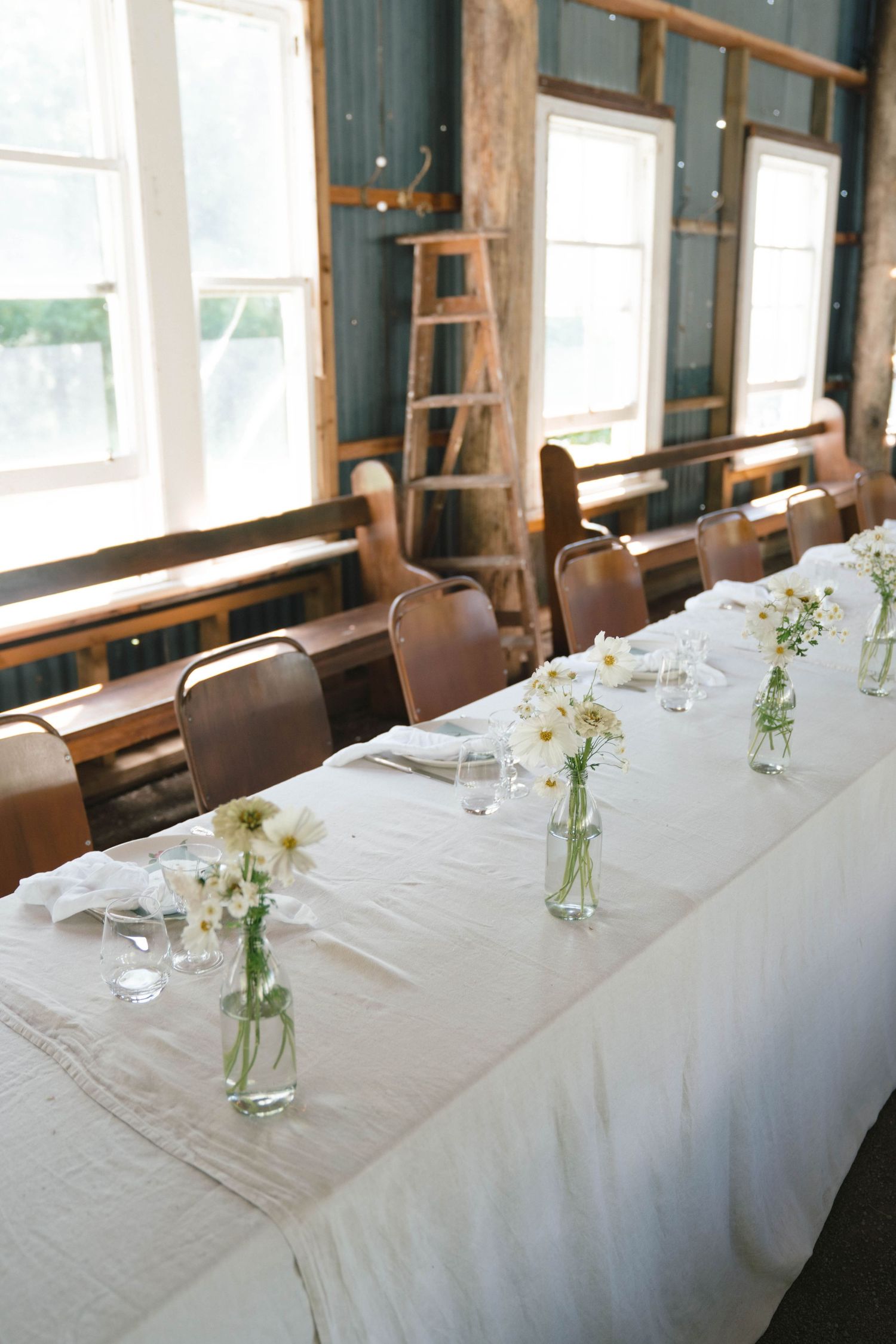 A long dining table adorned with white linens and small glass vases containing delicate white flowers in natural light.