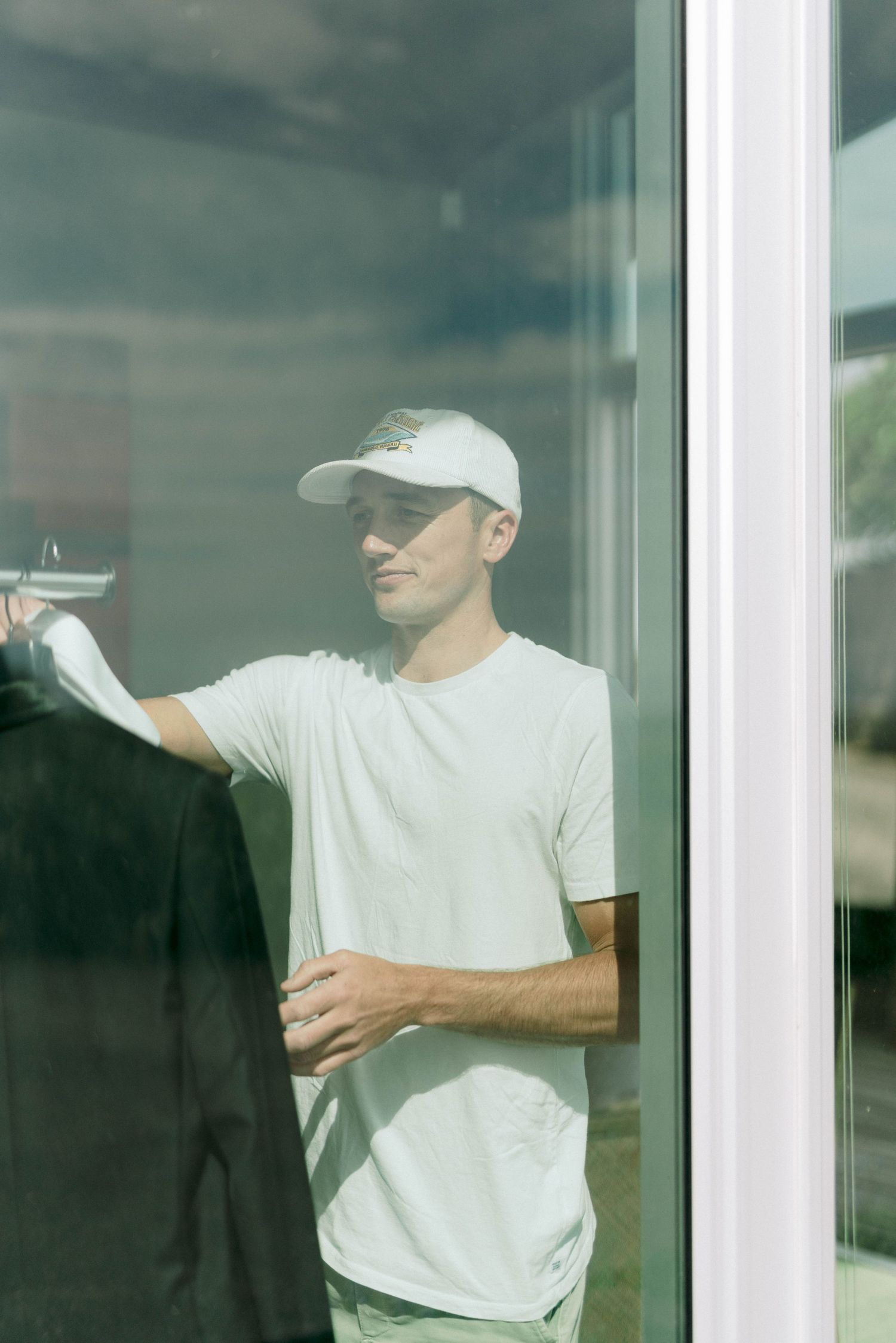 Person in white t-shirt and baseball cap checking formal wear through window reflection.