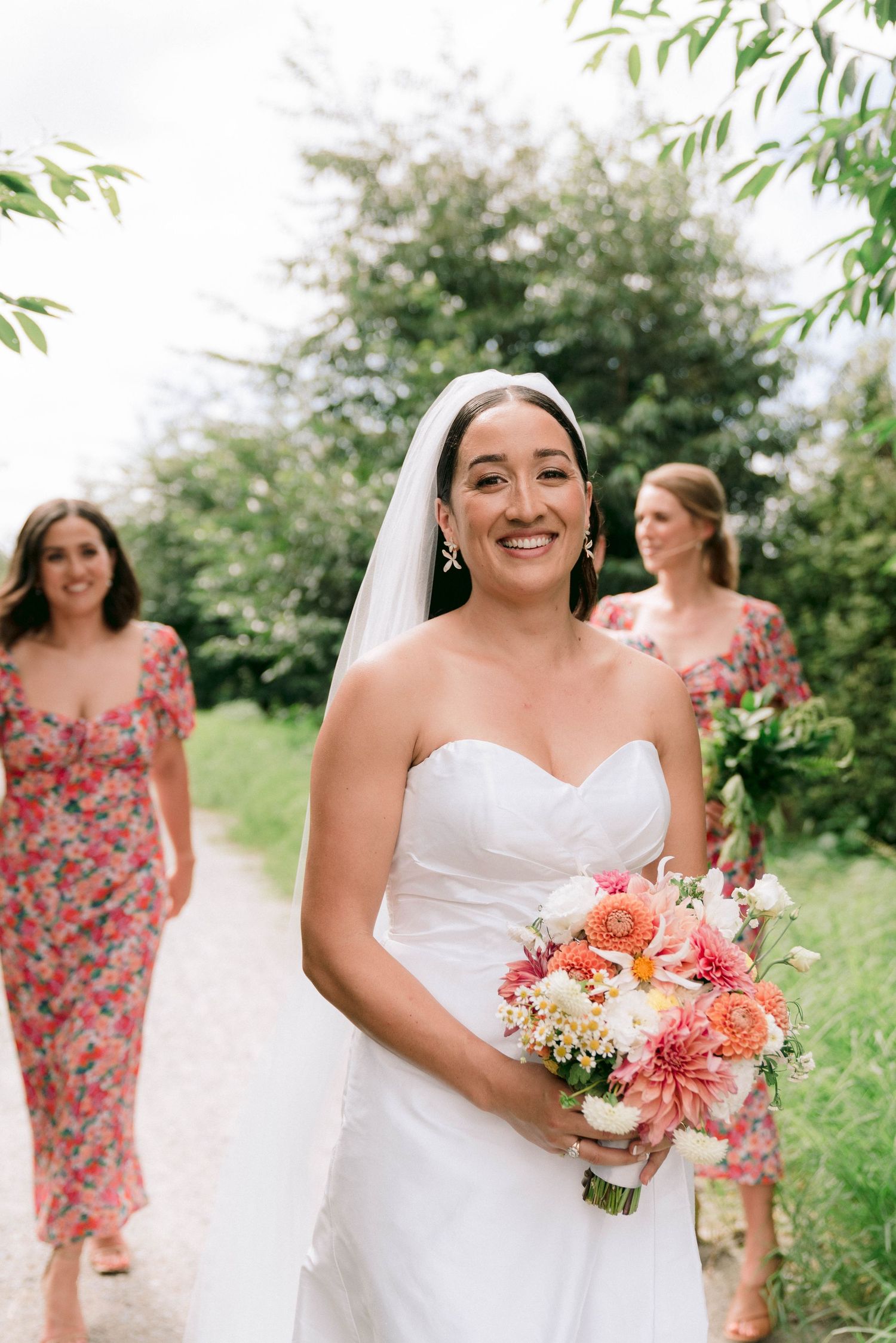 A bride in a white strapless gown holds a vibrant coral and pink bouquet while walking outdoors.