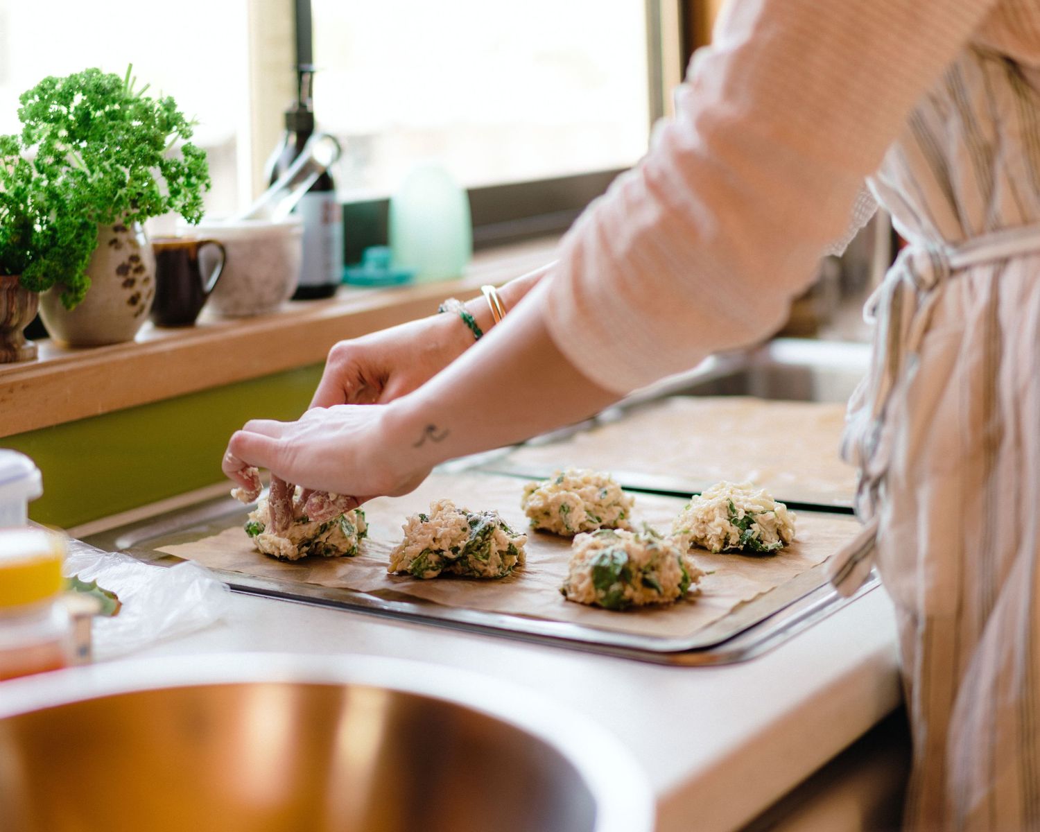 Hands placing homemade cookie dough scoops onto a baking sheet next to other kitchen items.