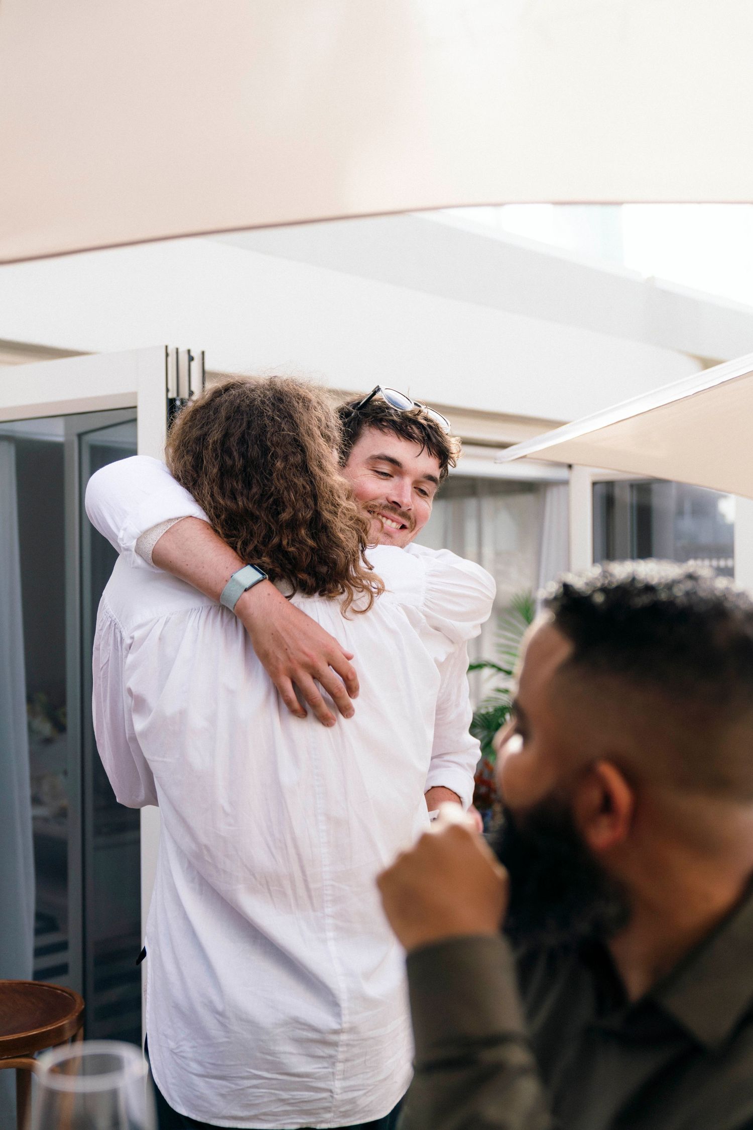 Two friends share a warm hug while another person watches at a casual indoor gathering.