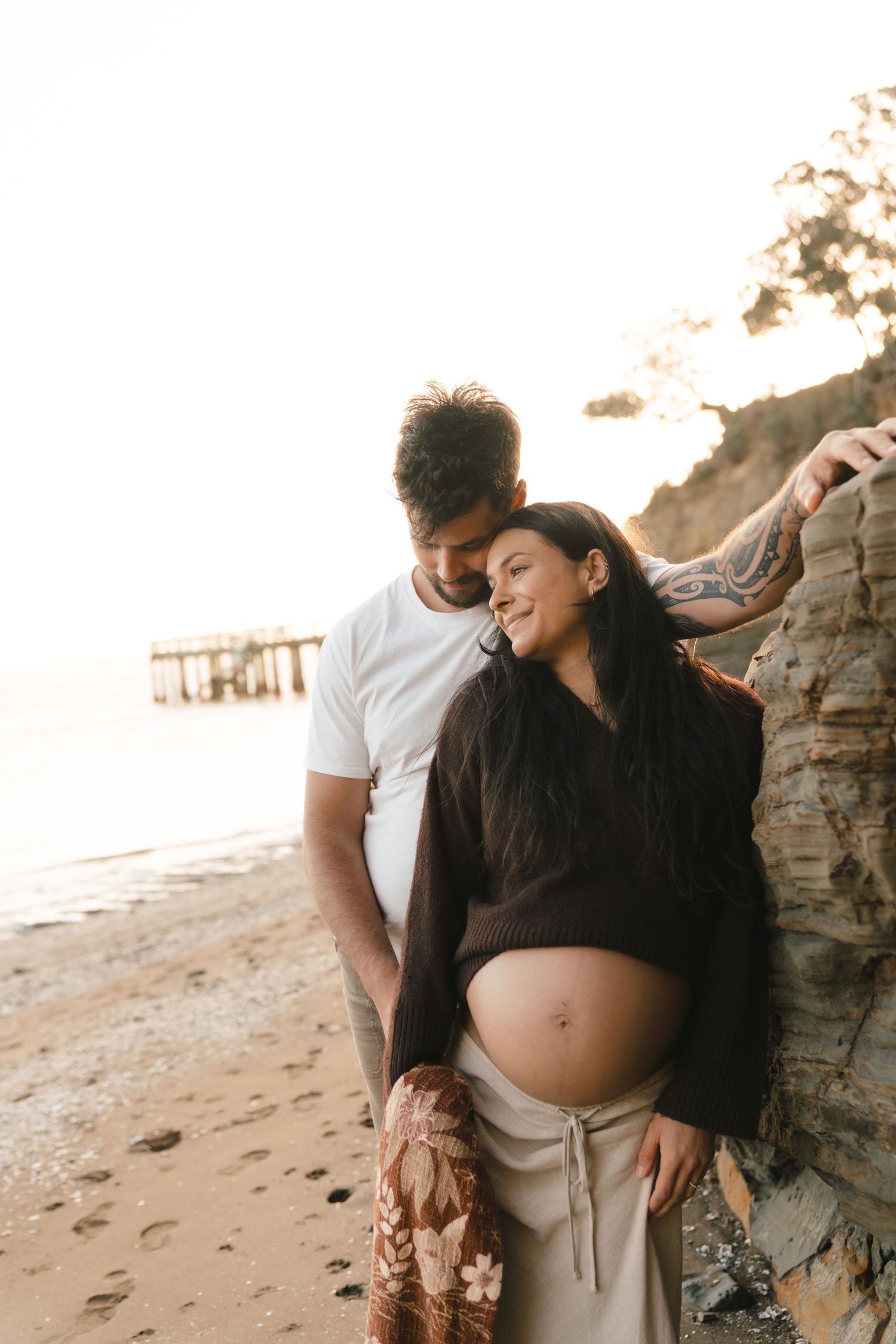 Expecting couple shares intimate moment on beach at sunset as mother shows off pregnant belly in brown sweater.