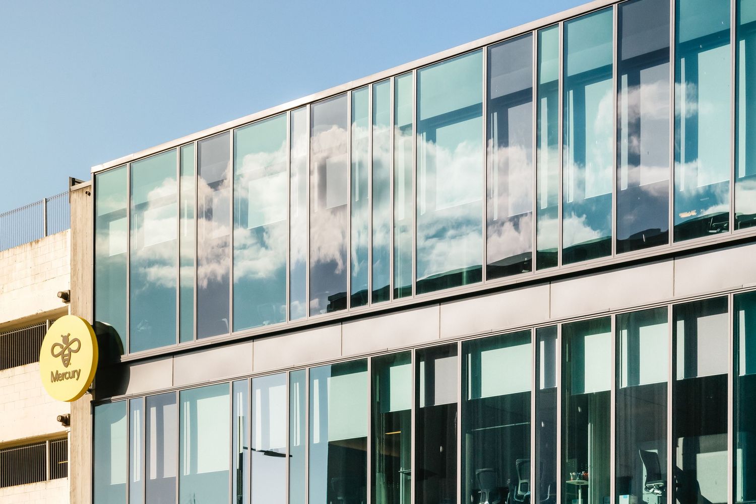 Modern glass office building exterior with rows of windows reflecting sky and clouds on a sunny day.