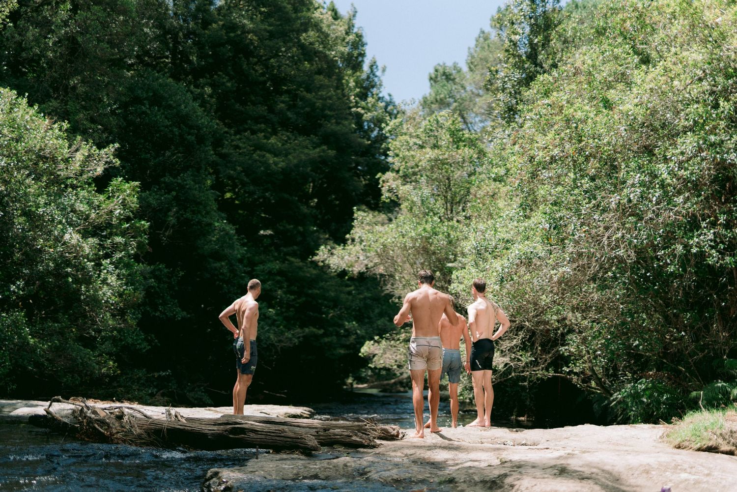 Three people enjoying a sunny day standing on rocks by a river in a forested area.