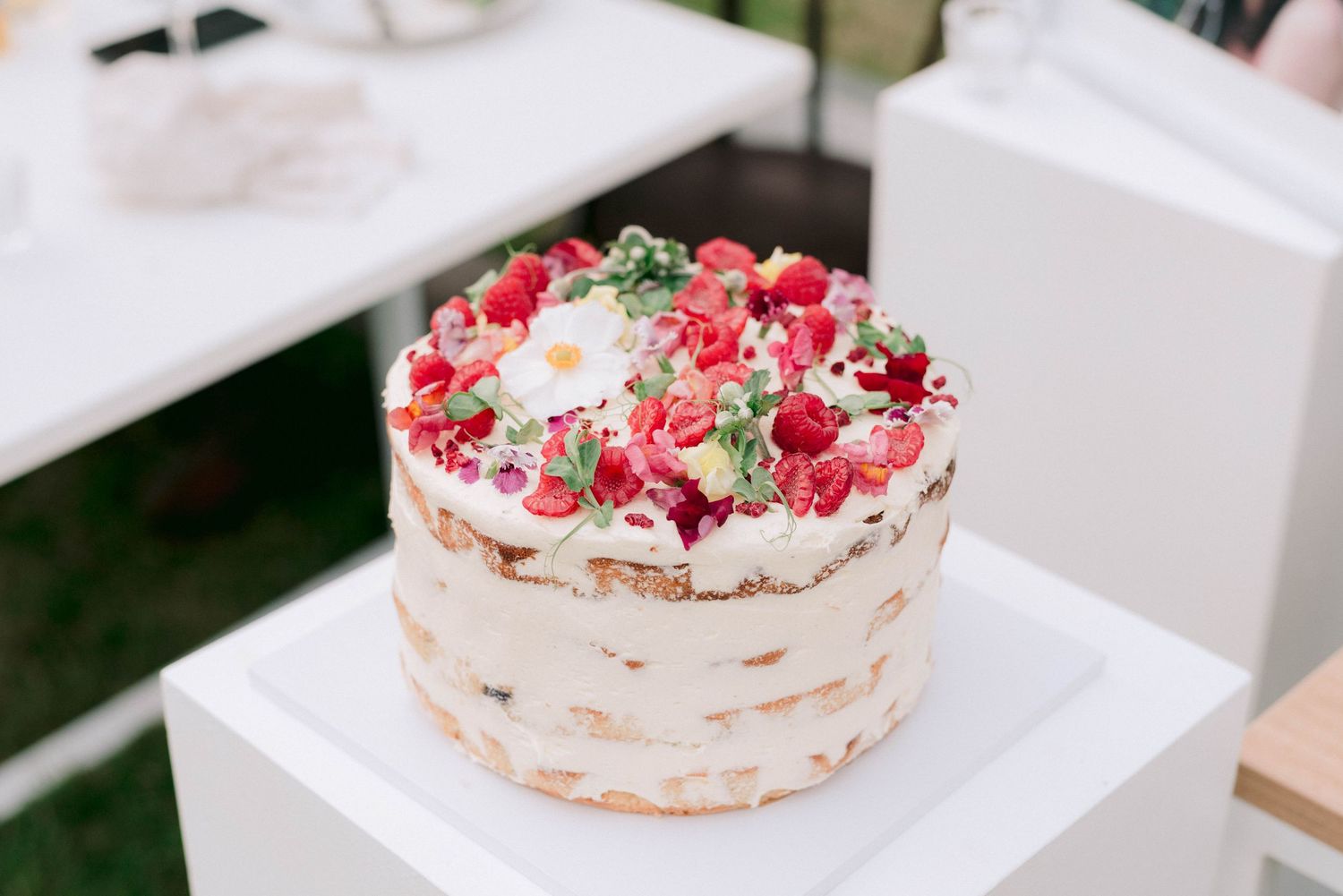 A layered cream cake decorated with fresh red berries and green herbs on a white stand.