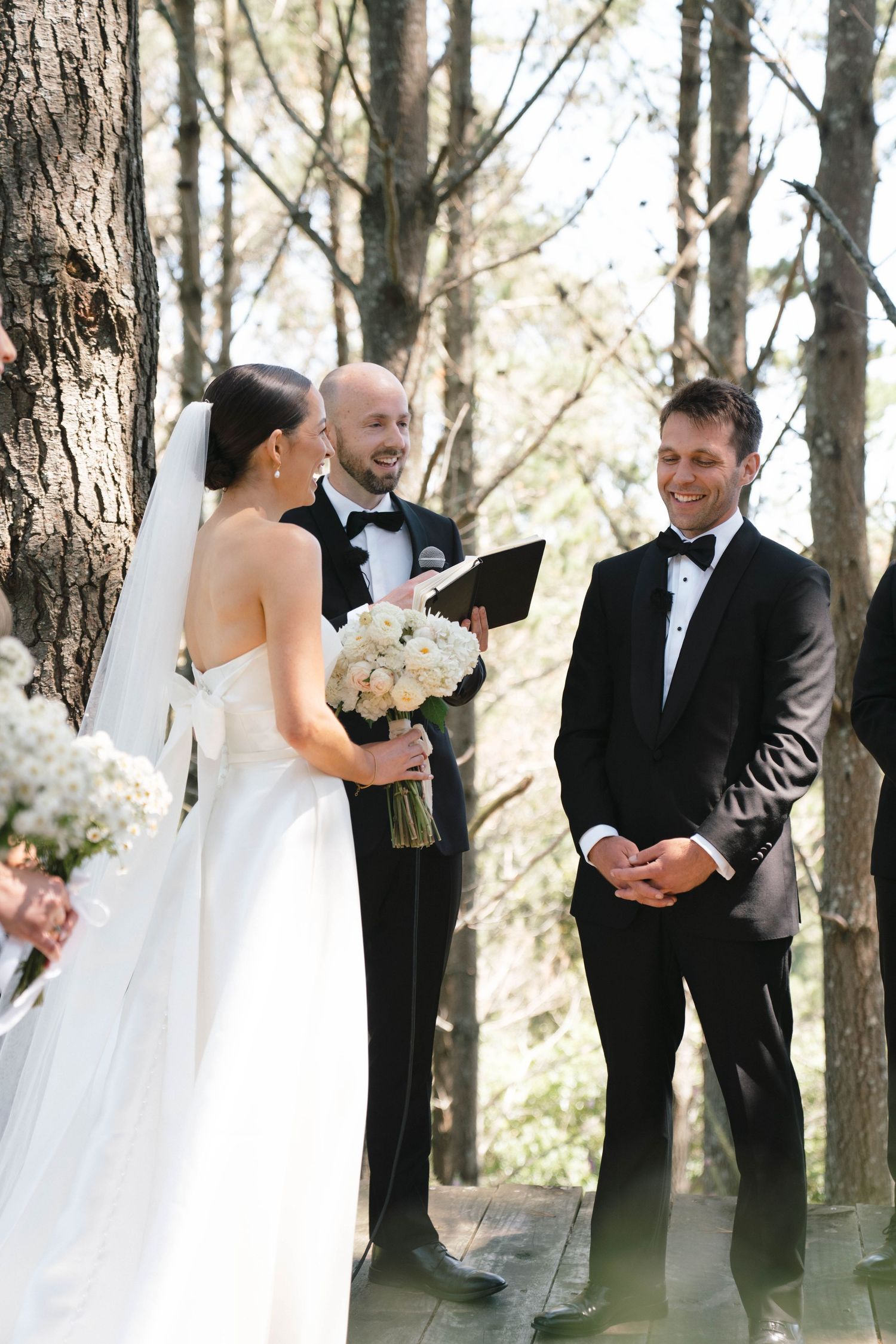 A wedding ceremony takes place in a serene forest setting with white floral arrangements and formal attire.