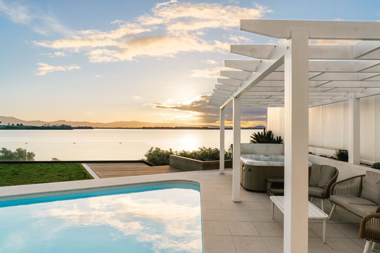 Luxurious waterfront patio with white pergola and infinity pool overlooking sunset view.