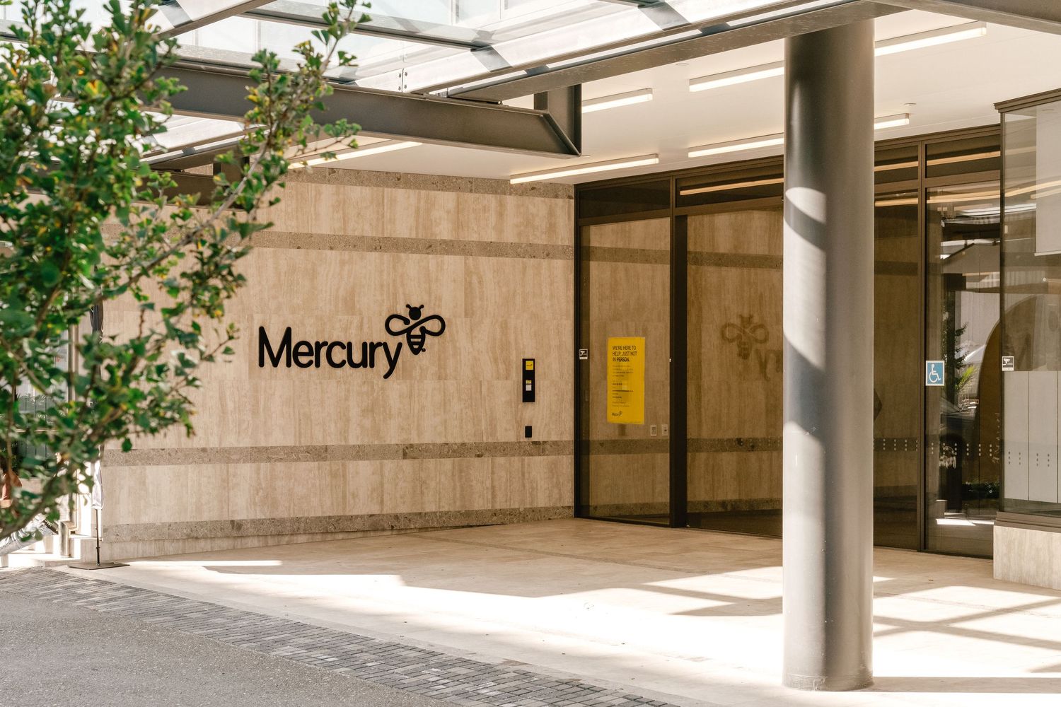 Modern office building entrance with Mercury signage and glass doors surrounded by concrete columns and green foliage.