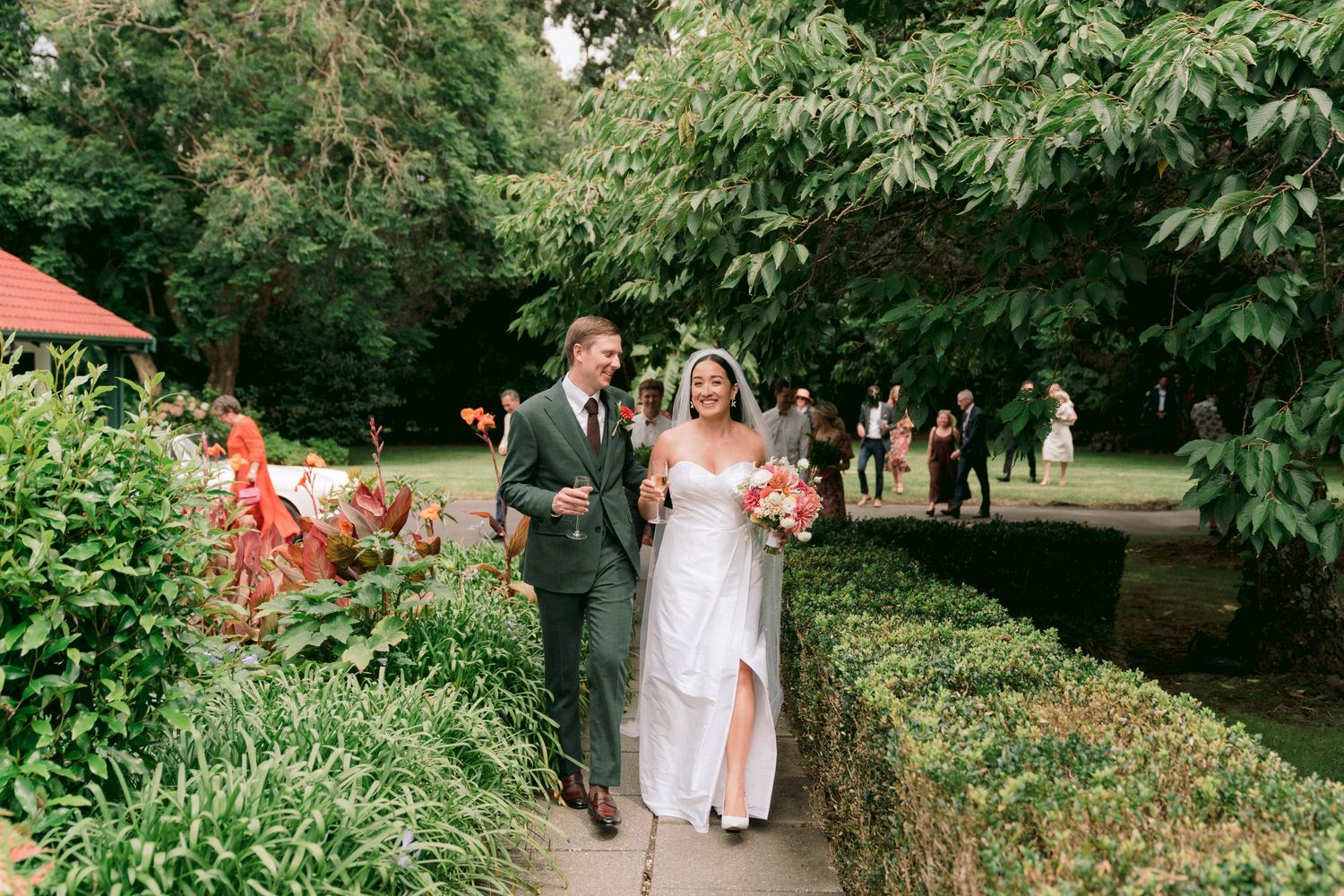 Wedding party walking through lush garden path surrounded by green foliage.