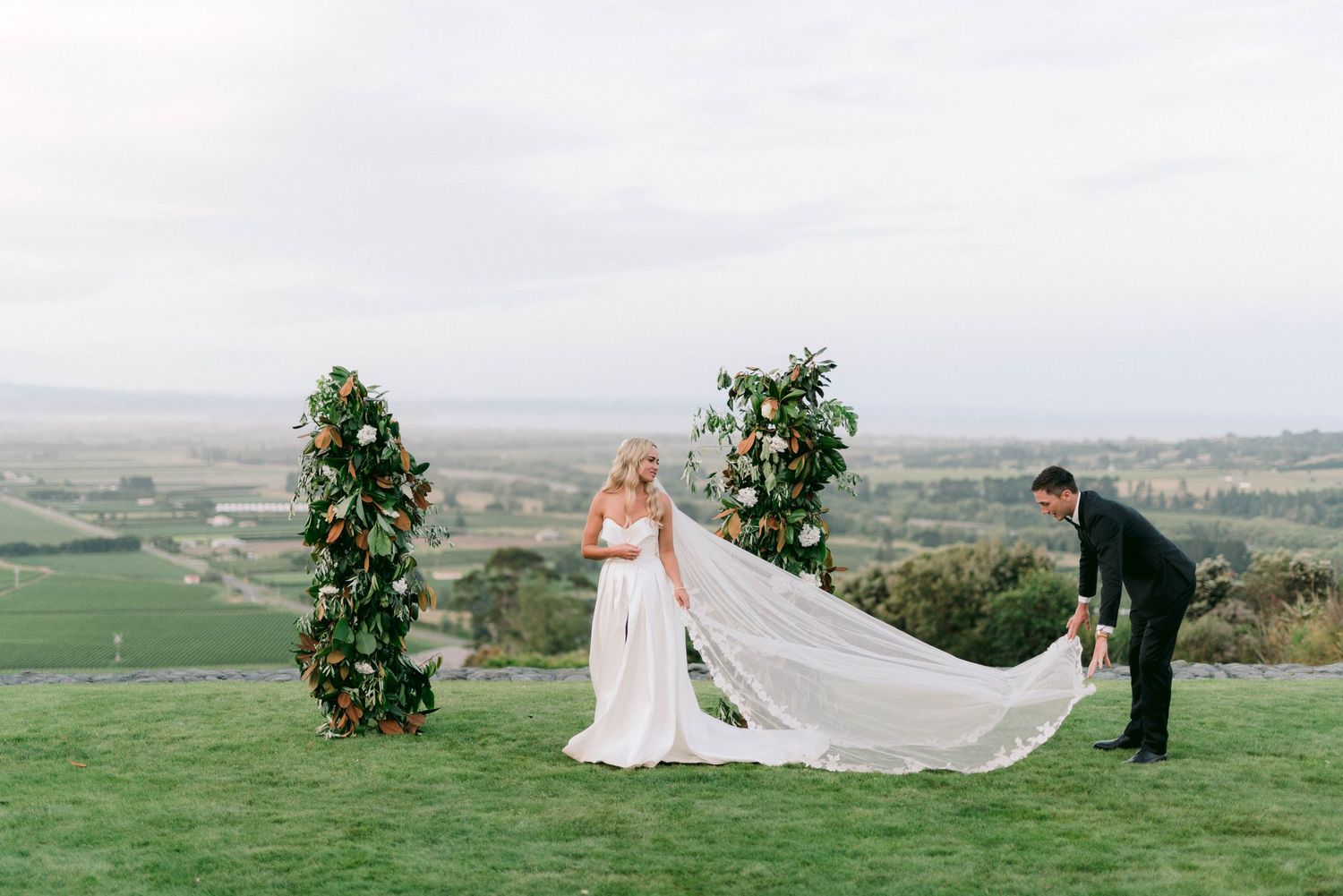 Scenic hilltop wedding ceremony setup with floral arch and flowing white veil against rolling countryside landscape.