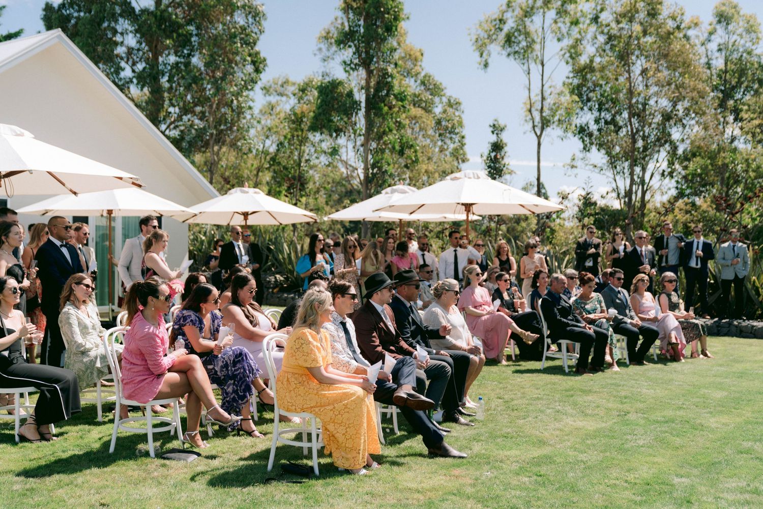 Wedding guests seated under white umbrellas on a manicured lawn during an outdoor ceremony.