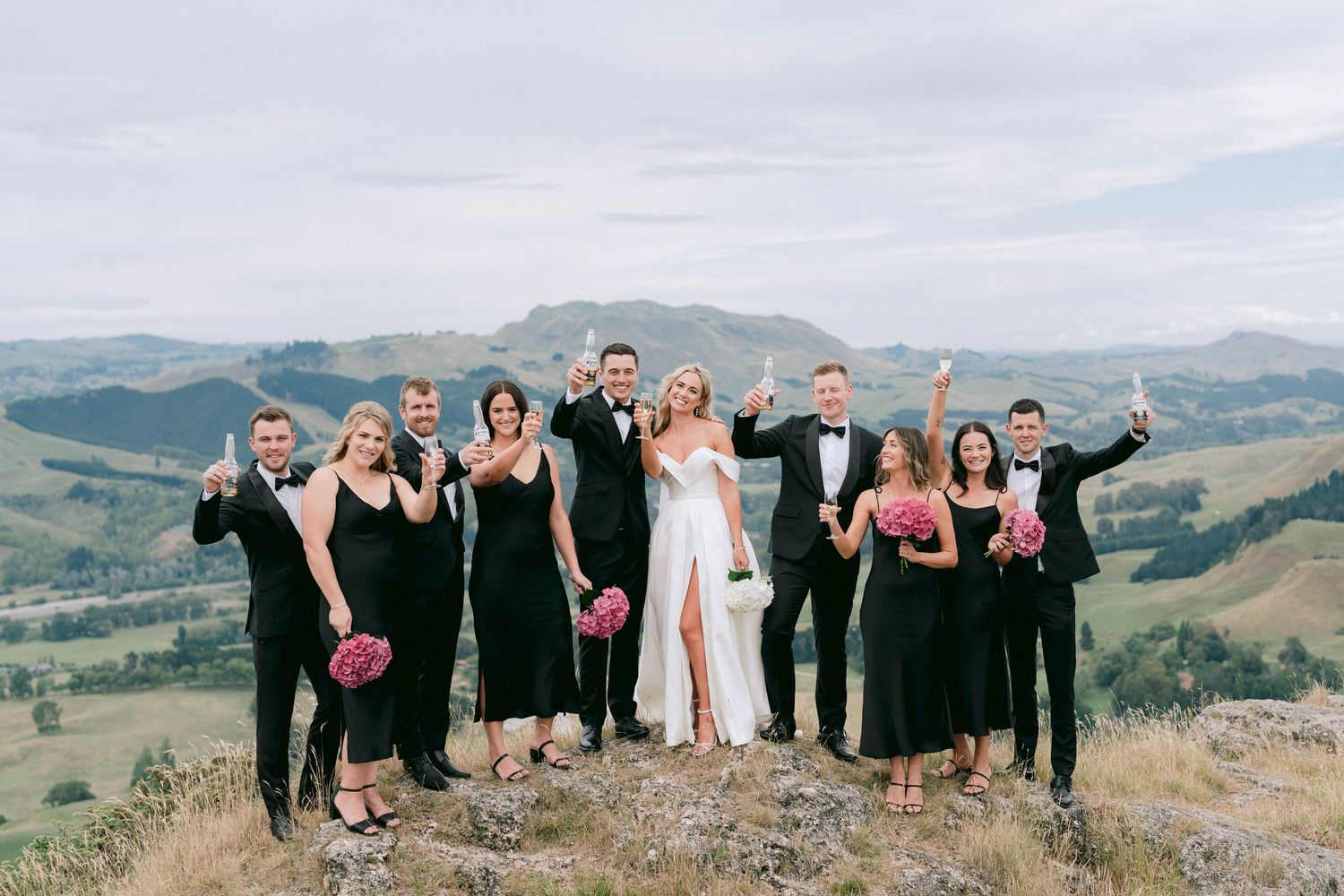 Wedding party in formal attire poses enthusiastically on a hilltop with mountain views.